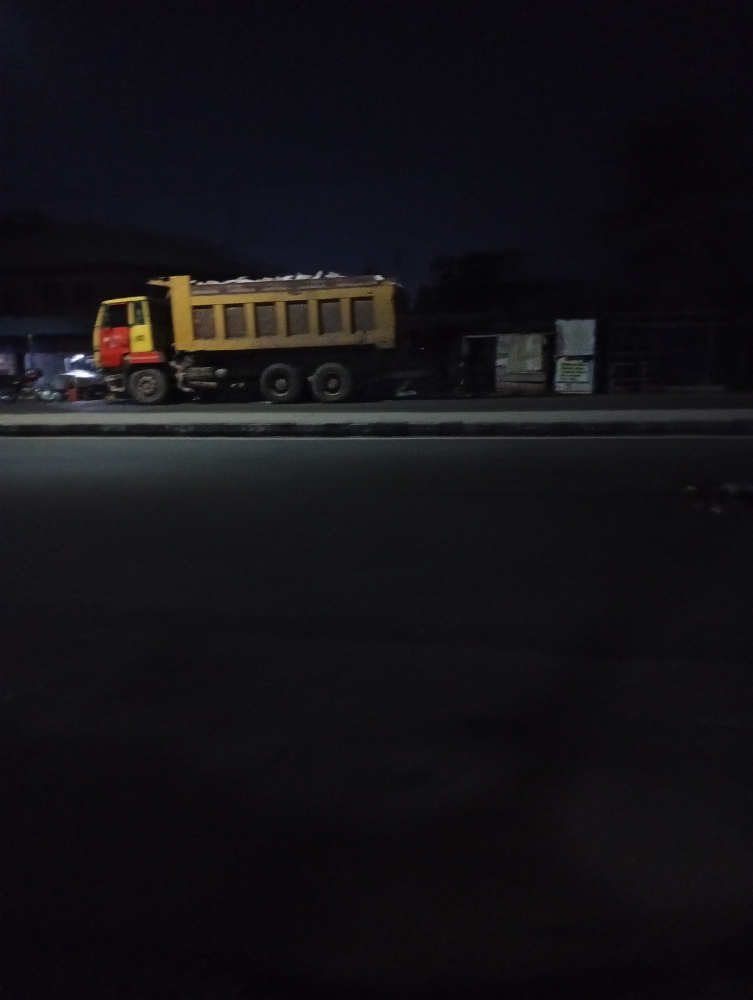 The image shows a yellow and red dump truck parked on a road at night in an urban environment. The truck bed appears to be loaded. There are structures and objects along the side of the road, likely shops or buildings. The road is dark, and the lighting suggests it is nighttime. The location is Unknown, Unknown.