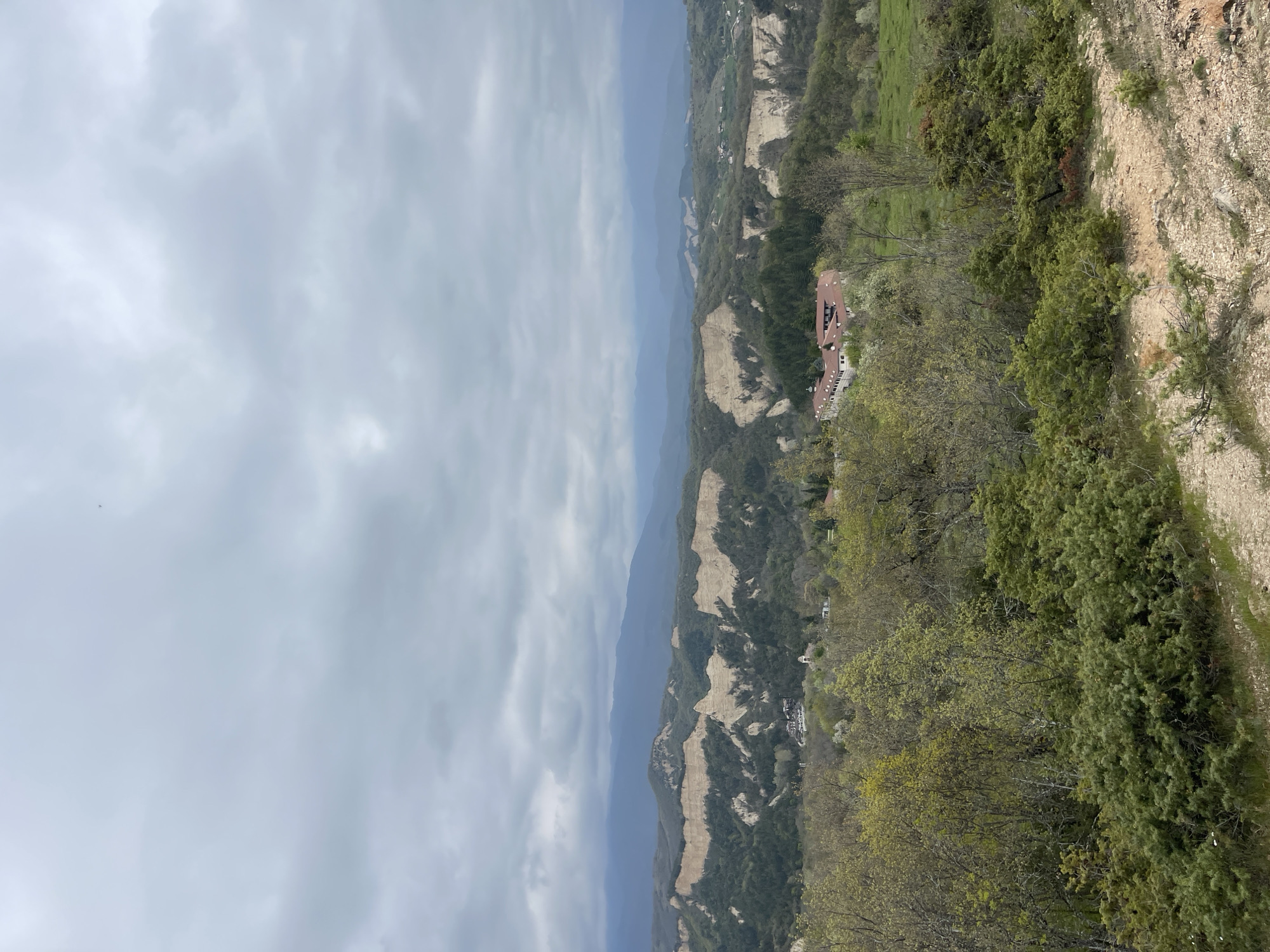 This is an outdoor, daytime shot of a mountainous landscape under a cloudy sky, likely taken from a high vantage point. The view consists of a deep valley with textured rocky terrain interspersed with green vegetation and trees. In the middle ground, there appears to be a building with a reddish-brown roof surrounded by more greenery. The terrain extends into the distance towards the right side of the frame. On the far left, the sky is overcast with a thick layer of clouds. The composition gives a sense of depth and scale to the landscape. The location appears to be Rozhen, Bulgaria, based on the terrain and vegetation.