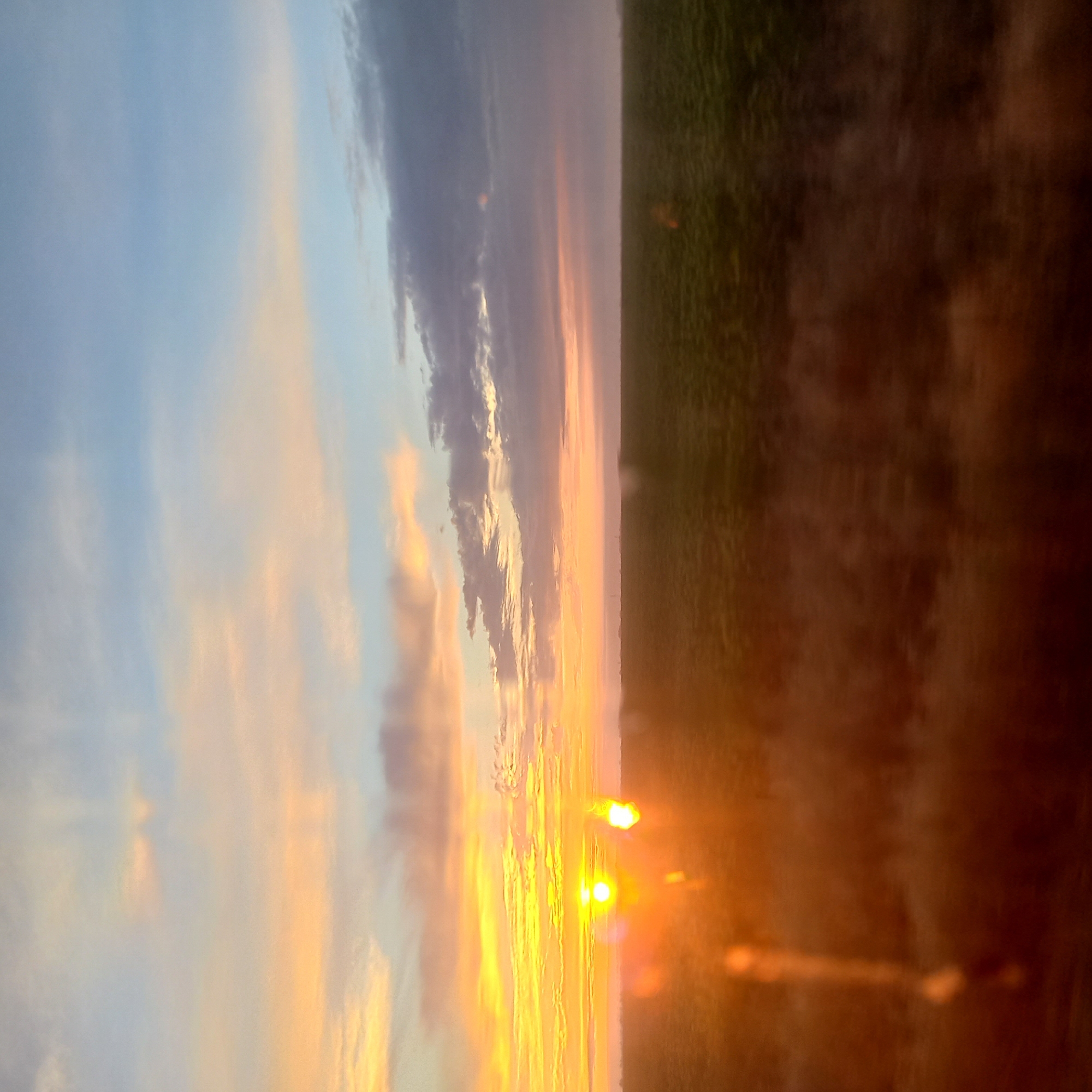 The image depicts a sunset over a flat, agricultural landscape. The sky is filled with clouds painted in hues of orange, yellow, pink, and blue. The sun, a bright orb, is visible near the horizon, casting a warm glow across the scene. In the foreground, a dark, blurred area suggests the presence of vegetation or a field, possibly with some dry grass or soil. The overall impression is one of a rural setting at dusk. The image does not contain any discernable features that would allow for specific identification of the location within Kropyvnytskyi, Ukraine.