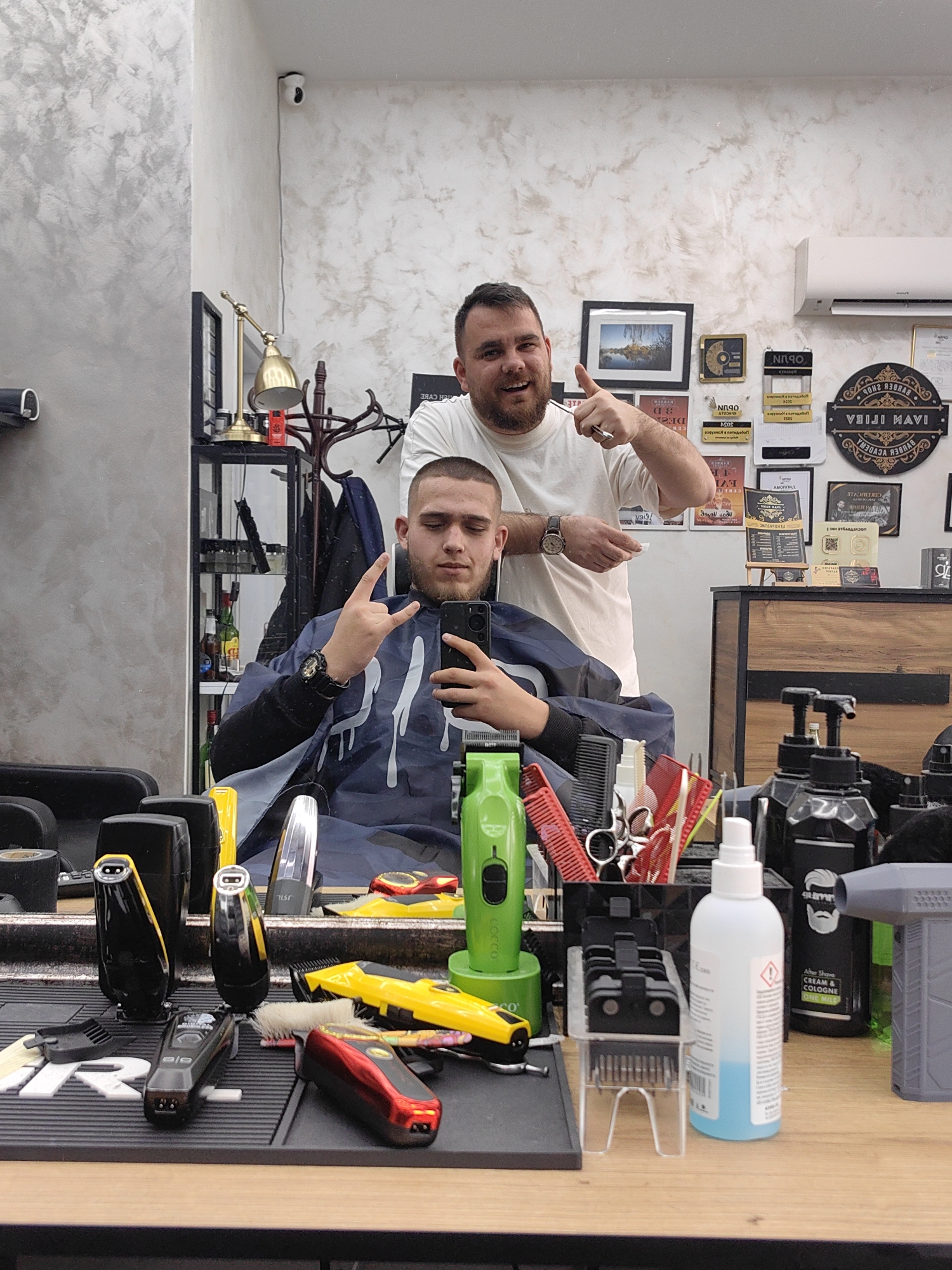 The image captures a scene inside a barbershop. A young man sits in the barber's chair, draped in a blue cape, holding a smartphone to take a selfie, in which he holds up a hand making the "rock on" gesture. Behind him, a barber stands and gives a thumbs-up.
In the foreground, a barber's workstation is visible. Various grooming tools and supplies are present, including clippers, combs, scissors, hair products, and a bottle of hand sanitizer.
The background features a decorated wall with framed pictures, signage, and a shelf with bottles and other items. A white air conditioning unit is mounted on the wall. A sign "VEIJI MAVI IVAN ILIEV BARBER ACADEMY" is also present.
Based on the signs and the style, it is likely the image was taken in a city in Bulgaria, most likely in Sofia.