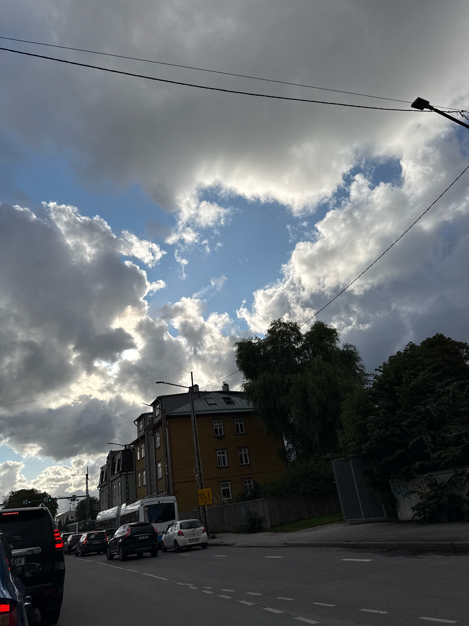The image captures a street scene in Tallinn, Estonia, under a dramatic sky filled with dark, volumetric clouds and patches of blue. A traffic jam is visible on a paved road in the foreground, with several cars, including a black SUV, a white hatchback, and a white bus or tram. The buildings lining the street are primarily residential, with a prominent yellow wooden structure featuring multiple windows. Trees with dark green foliage are present on the right side of the frame, casting shadows. Power lines are strung across the sky. A street lamp is visible on the right. The scene suggests a cloudy day with limited sunlight.
