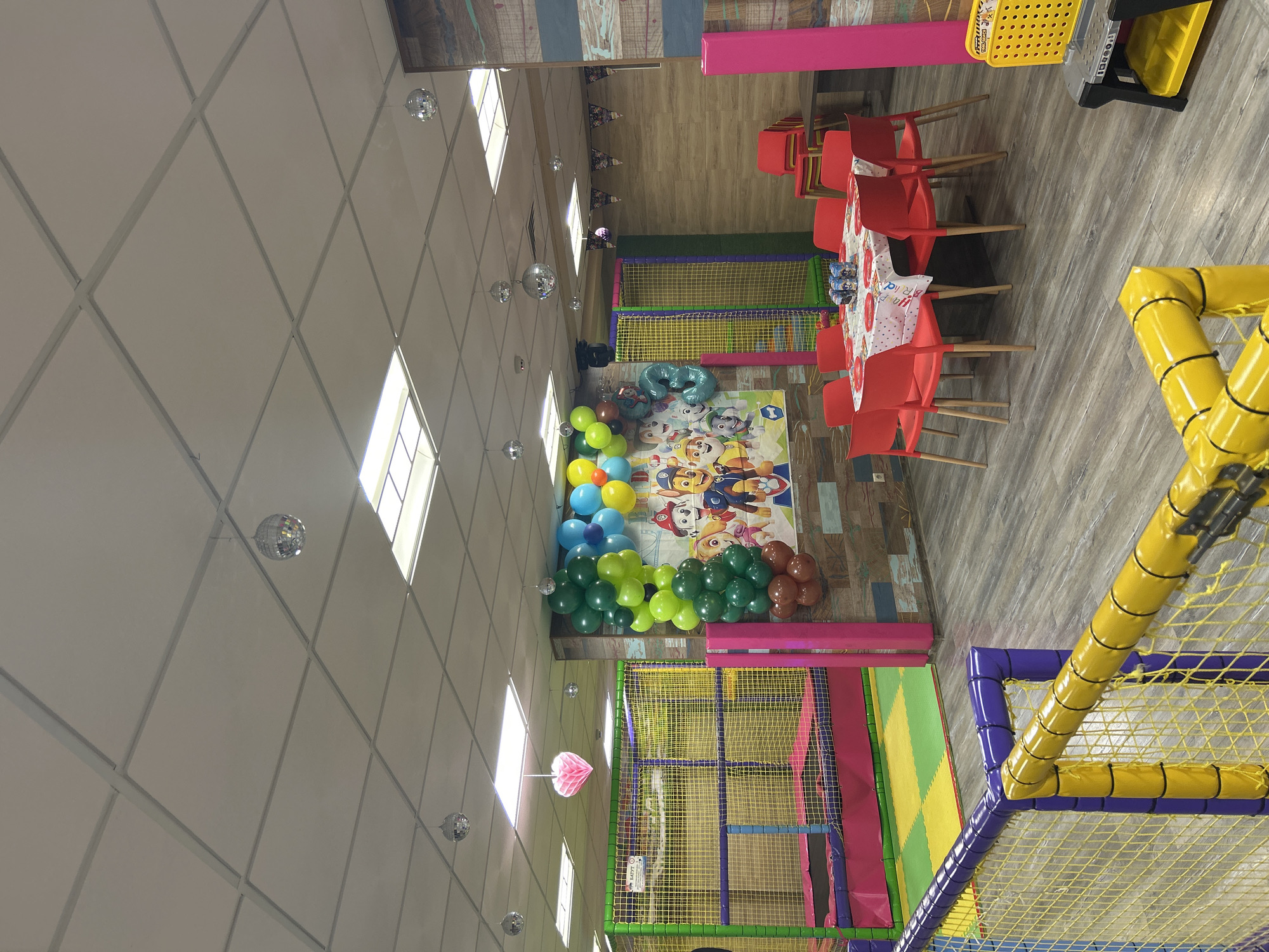 The image shows the interior of a brightly colored indoor playground, possibly located in Plovdiv, Bulgaria. The space is decorated for a children's birthday party, evidenced by the colorful balloons, a Paw Patrol-themed backdrop, and a table set for a celebration with red chairs. Playground equipment with yellow, purple, and green elements is visible. The ceiling is white and features recessed lighting and disco balls. The floor is wood-patterned. There are no people visible in the frame.