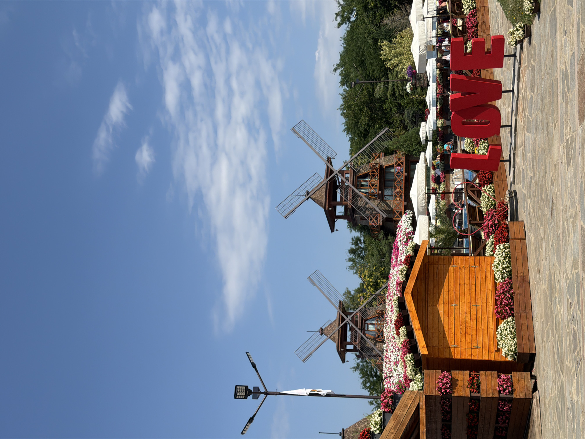 The image depicts an outdoor scene in the city of Gorna Malina, Bulgaria, likely a park or a recreational area. In the foreground, a large red three-dimensional "LOVE" sign stands on a paved stone ground. To the left of the sign, a small wooden structure with a pitched roof is adorned with planters filled with white and pink flowers. Behind this, a more elaborate wooden structure with planters filled with a profusion of pink and white flowers stretches across the frame. Further in the background, two rustic wooden windmills with visible blades rise above a lush green treeline. A modern street lamp with multiple LED lights is visible to the left of the windmills. White umbrellas, possibly for a seating area, are also scattered throughout the mid-ground, with some patrons visible seated beneath them. The sky above is a clear blue with sparse white clouds.