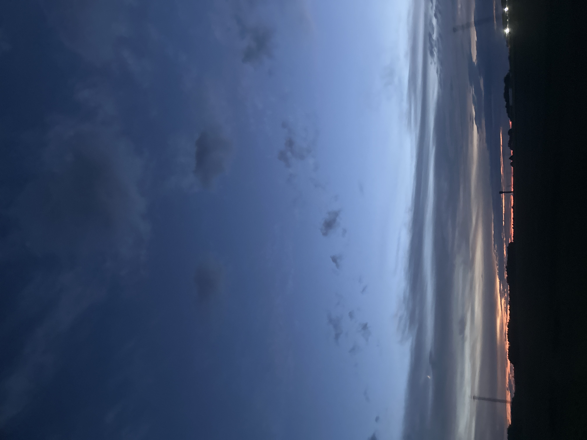 The image shows a dark silhouette of trees and a field against a twilight sky over the Грозаўскі сельскі Савет, Belarus. The sky transitions from a deep blue at the top to lighter shades of blue and orange near the horizon, with scattered clouds. There are faint lights visible on the right side of the horizon, possibly from buildings or streetlights. A thin crescent moon is visible in the sky above the horizon on the left.