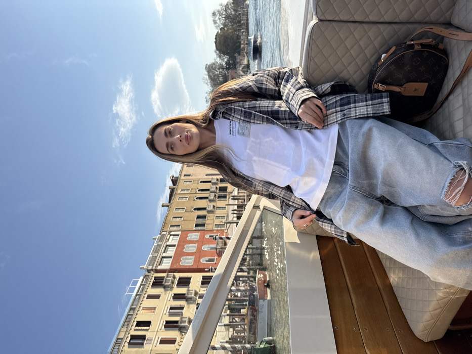 A female subject is seated on a water taxi in a canal in Venice, Italy. The subject has long, light brown hair and is wearing a white t-shirt with a small graphic, an open grey and white plaid long-sleeve shirt, and light blue ripped jeans, revealing her right knee. She is looking slightly upward and to her right with a neutral expression. A brown Louis Vuitton monogram handbag rests on the quilted cream-colored seat to her right. The boat features wooden paneling and a white railing with glass inserts. In the background, a row of multi-story, historic Venetian buildings with yellow, red, and beige facades lines the canal. Several other boats are visible on the sun-dappled water. The scene is illuminated by bright sunlight under a clear blue sky with a few faint clouds.