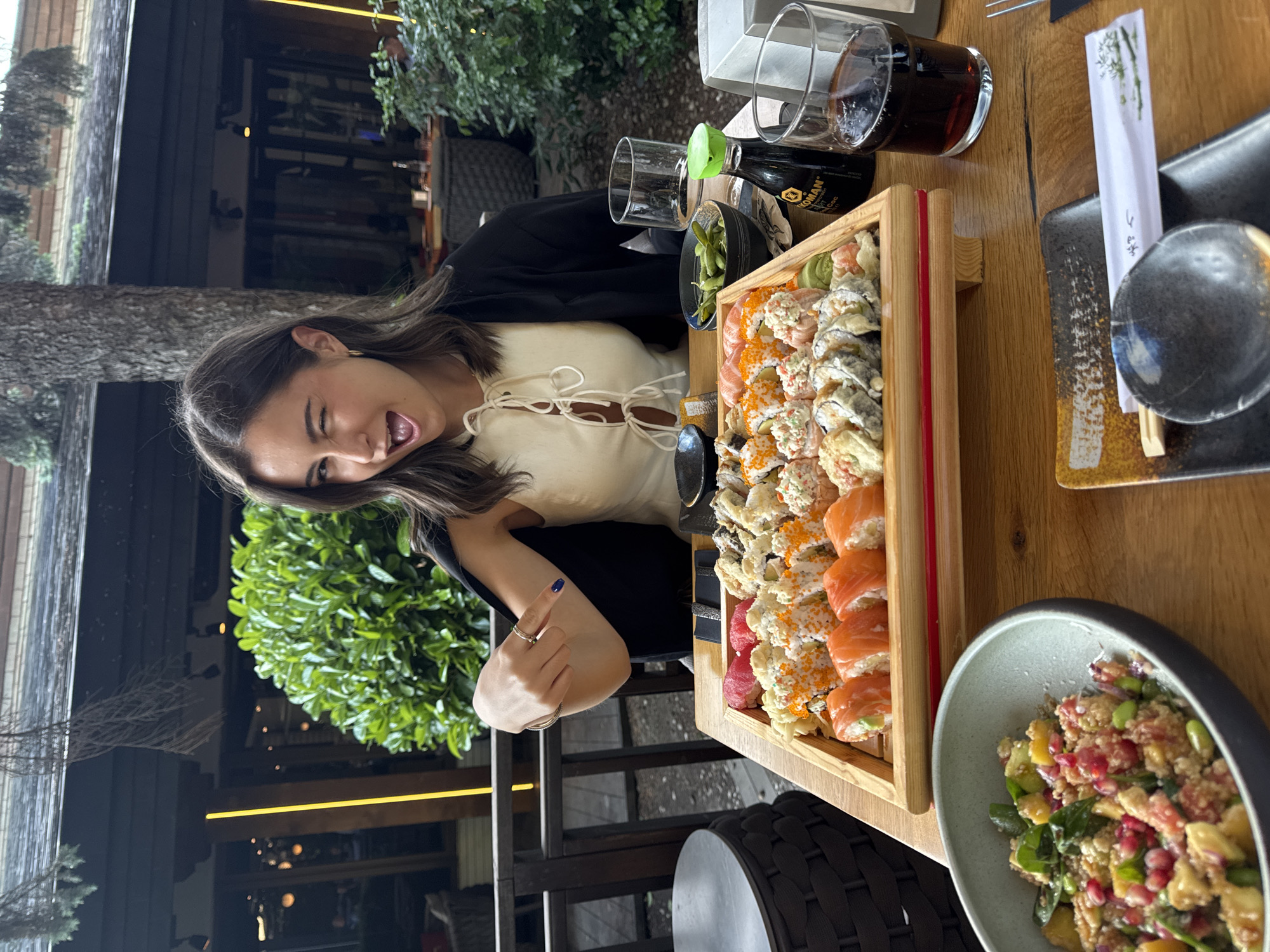 The photograph shows a young woman seated at a wooden table covered in sushi and side dishes. The sushi includes salmon, tuna, and California rolls. There are also edamame beans, salad, soy sauce, and a glass of dark soda. The woman is winking and giving a thumbs-up gesture. The environment appears to be a restaurant with outdoor seating. Based on the style of the buildings and vegetation visible in the background, this location is likely in Sofia, Bulgaria.