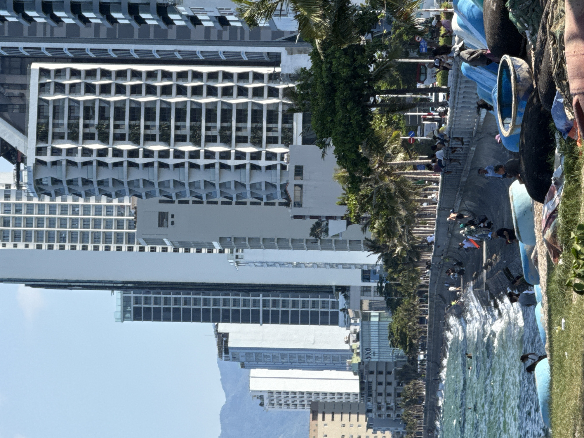 This media file captures a vibrant scene along the coast of Bac Nha Trang Ward, Vietnam. The image features a row of modern, multi-story buildings with striking geometric facades, interspersed with lush palm trees. In the foreground, the turquoise water of the sea churns with visible waves, suggesting a breezy day.
Along the waterfront, a concrete promenade is bustling with activity. Numerous people are gathered, some sitting, others standing, and a few appear to be walking. They are dressed casually, indicating a warm, sunny day, possibly mid-morning or afternoon, given the bright lighting and clear sky. The presence of boats, some with distinctive blue hulls, along the edge of the water suggests a maritime setting, perhaps a harbor or a popular tourist area. The overall mood seems relaxed and lively, with a clear sense of community and outdoor enjoyment.
A notable detail is the presence of various objects on the promenade, including what appear to be large, dark-colored cylindrical objects, possibly buoys or floats, along with fabric items. A small, circular blue opening is visible within one of these objects. A glimpse of text is visible on one of the buildings; "YOLO" is clearly legible. The scene combines urban architecture with natural coastal elements and human activity, painting a picture of a lively day in a scenic Vietnamese location.
