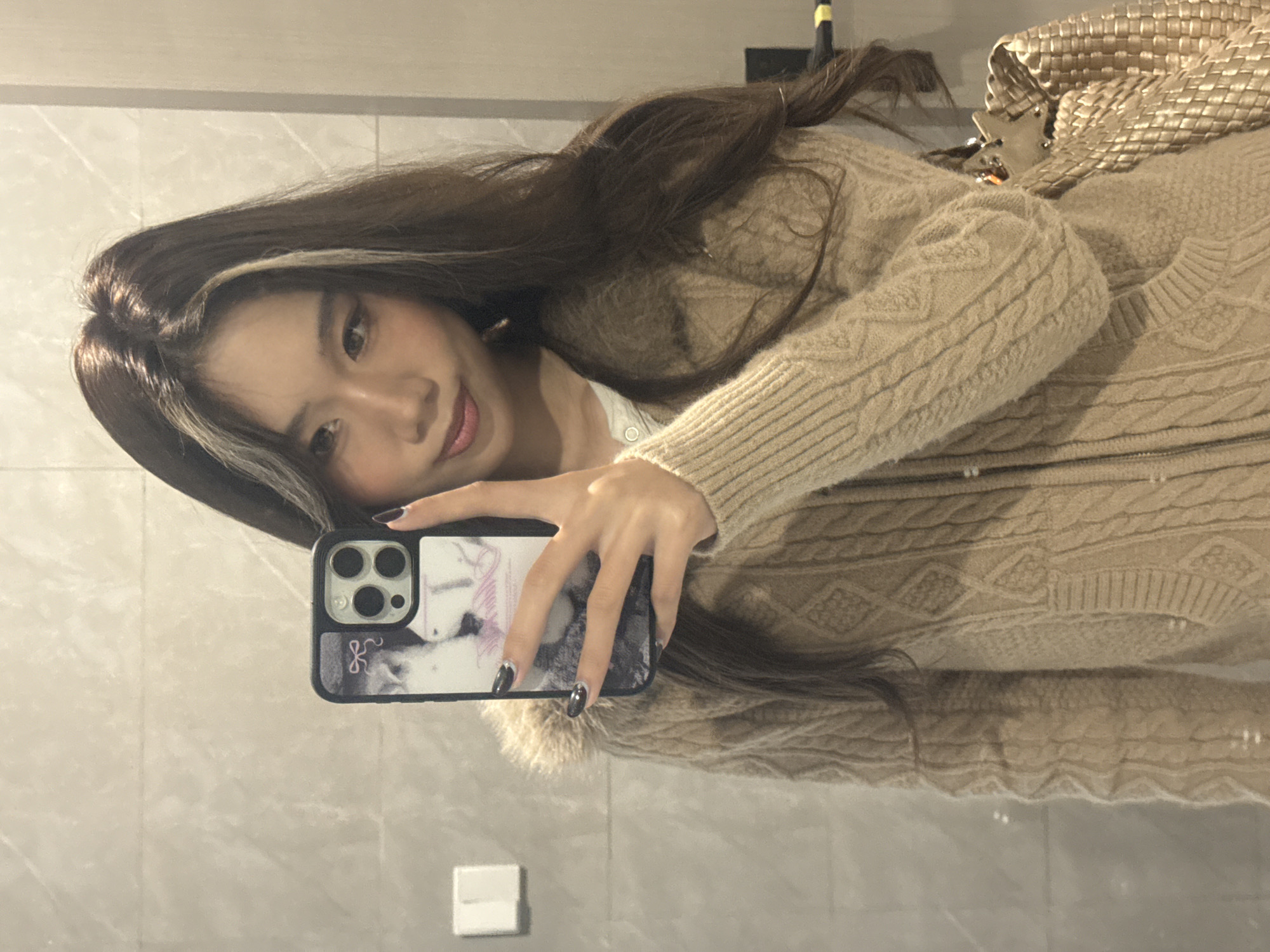 A female individual is depicted taking a mirror selfie in an indoor setting, identified as Nan'an District, China. The subject has long, dark brown hair with two distinct light-colored streaks framing the face. She is wearing a beige cable-knit cardigan with visible fur trim around the collar or hood, and a light-colored top beneath. Her nails are manicured with a dark polish that transitions to a lighter, possibly glittered, tip.
She holds a dark-colored smartphone, likely an iPhone with a prominent triple-camera array, in her right hand, positioning it to capture her reflection. The phone case features a graphic design, appearing to show two light-colored felines or similar animals, with faint pink text or a ribbon detail. A light-colored woven bag is visible on her right side, hanging from her shoulder or arm.
The environment reflected in the mirror consists of a light grey or beige tiled wall, with horizontal grout lines visible. A white rectangular light switch or electrical outlet is present on the left side of the reflection. On the right, a vertical light-colored fixture, possibly a door frame or partition, is partially visible, featuring a yellow and black striped band near its base. The lighting is uniform, suggesting an interior space.