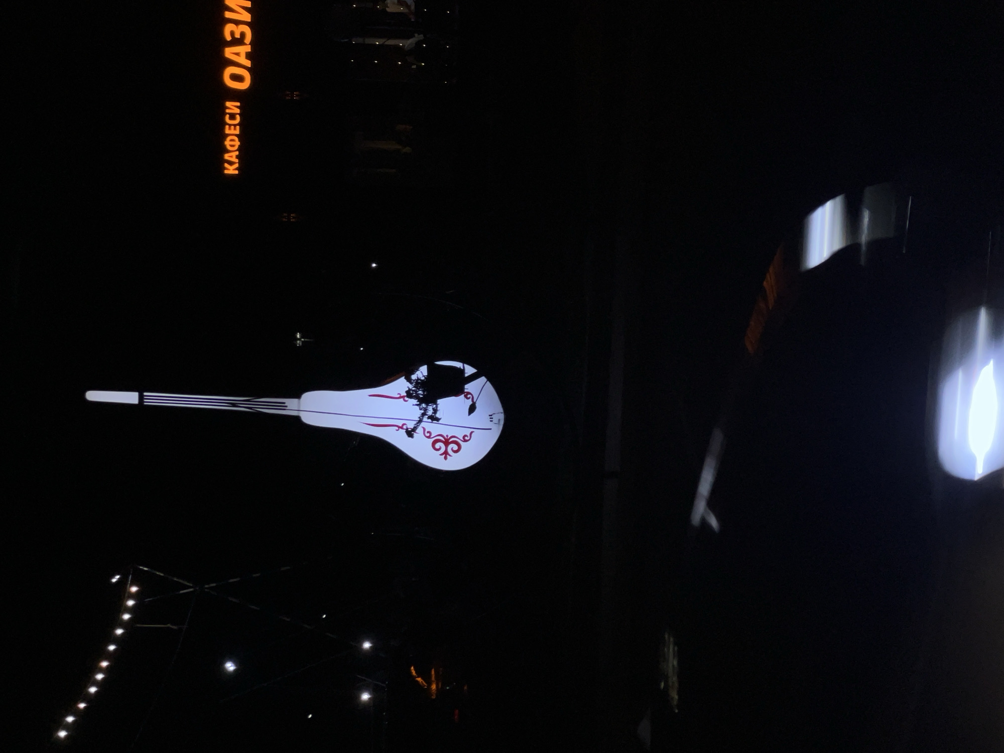 An illuminated, bulb-shaped installation resembling a musical instrument stands against a dark night sky. The installation features a white surface with red decorative elements and a silhouette of a person with a musical instrument. In the background, a neon sign reads "КАФЕСИ ОАЗИ" in orange lettering. String lights are visible in the upper left corner. The ground below the installation is dark and reflects some light. This scene is located in Ak-Suu, Kyrgyzstan.