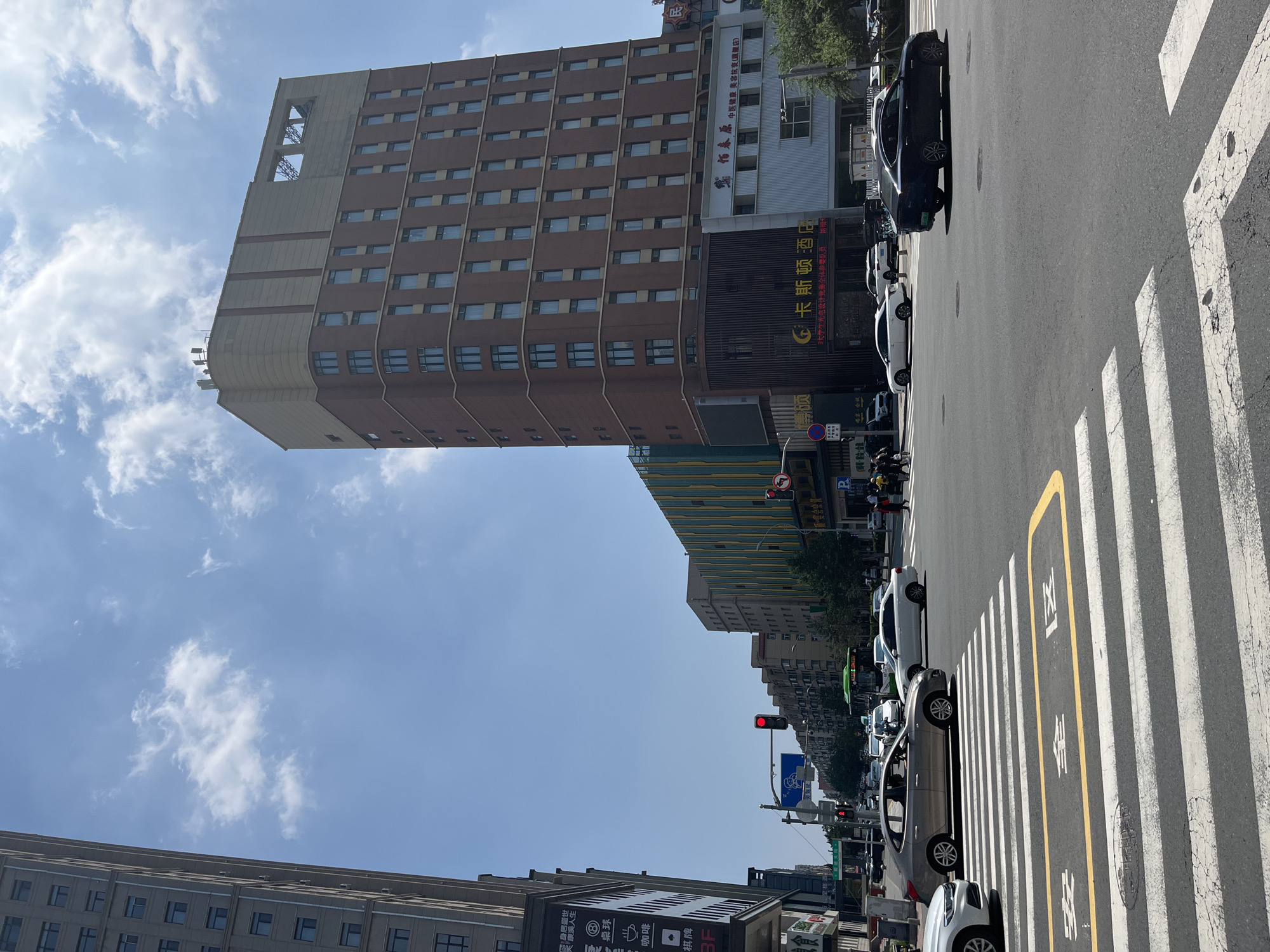 In Changchun City, China, on a bright, clear day, a busy intersection is captured with traffic flowing under a blue sky dotted with white clouds. A tall, modern building with a brown brick facade and a beige top section dominates the right side of the frame. The building features numerous rectangular windows. In front of it, a white structure with blue lettering and a red awning is visible, likely a shop or restaurant. To the left of this main building, a narrower, grey building stretches upwards.
The intersection itself is marked with clear white pedestrian crosswalks and yellow markings on the asphalt. Several cars are present: a silver sedan is stopped at the crosswalk, a white sedan is in the background, and a black sedan is moving through the intersection. A green bus is also visible, traveling down the street. Traffic lights show a red light for one direction. Pedestrians are seen crossing the street on the right side of the image. A large banner with Chinese characters is affixed to the side of the building on the left. The environment appears to be a commercial district with multiple stories of buildings lining the streets.