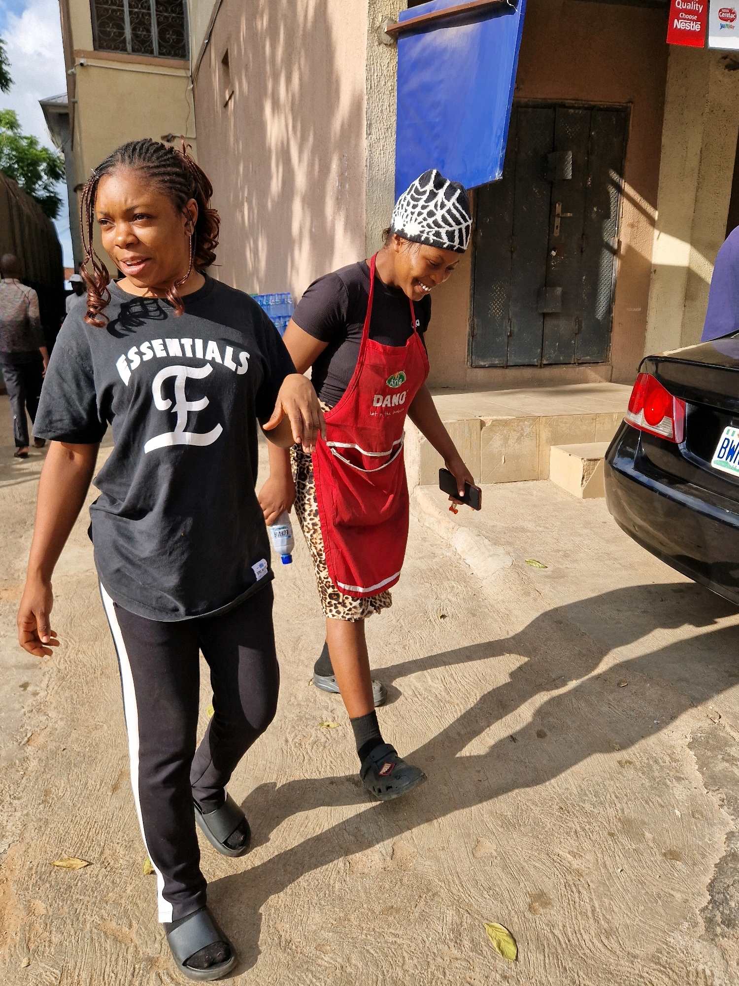 Two women are walking outdoors in what appears to be an urban or semi-urban environment. The woman on the left is wearing a black t-shirt with white lettering that reads "ESSENTIALS" and a large stylized "E" emblem. She also has black pants with a white stripe down the side and black slip-on sandals. Her hair is styled in braids with some curly extensions. The woman on the right is wearing a black t-shirt, a red apron with "DANO" printed on it, and a leopard-print skirt. She has a black and white web-patterned head covering. She is holding a black smartphone in her right hand. A black car is visible to the right of the frame. The ground is paved with concrete and shows signs of wear. There are shadows cast by the people and objects, indicating it is daytime. The background includes a building with a tan facade, a closed metal door, and some signage. There are also some cases of bottled water stacked near the building.