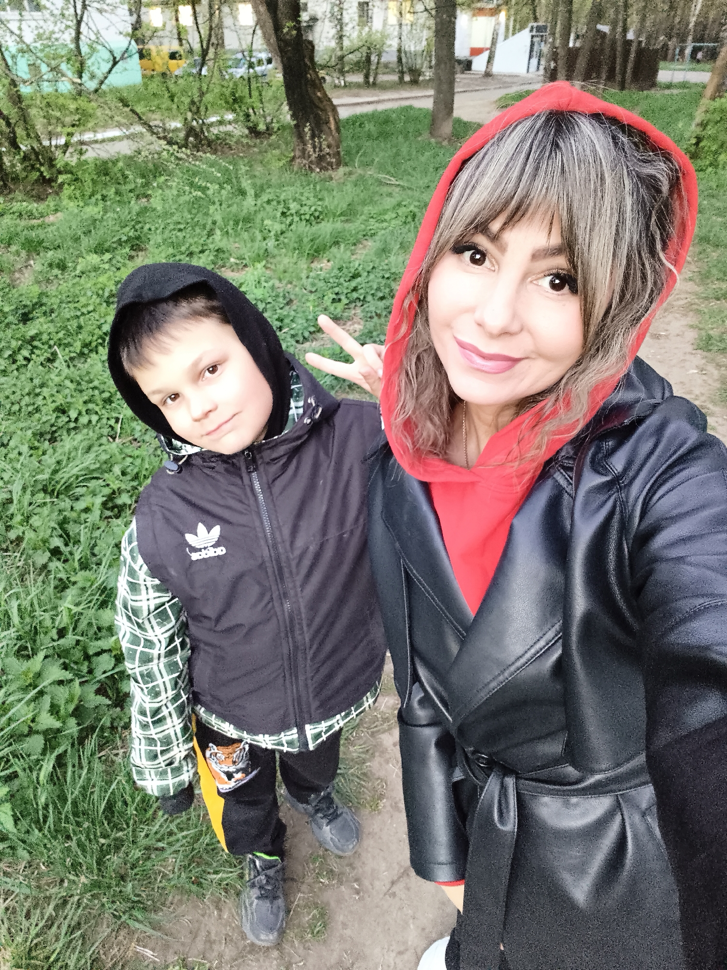 A selfie of a woman and a boy outdoors. The woman is on the right, wearing a black leather jacket over a red hooded sweatshirt. The boy is on the left, wearing a black hooded vest with an Adidas logo, and pants with a tiger image. He holds up a peace sign. They stand on a dirt path with green foliage. The location is likely Chaikovsky, Russia. Trees and buildings are in the background.