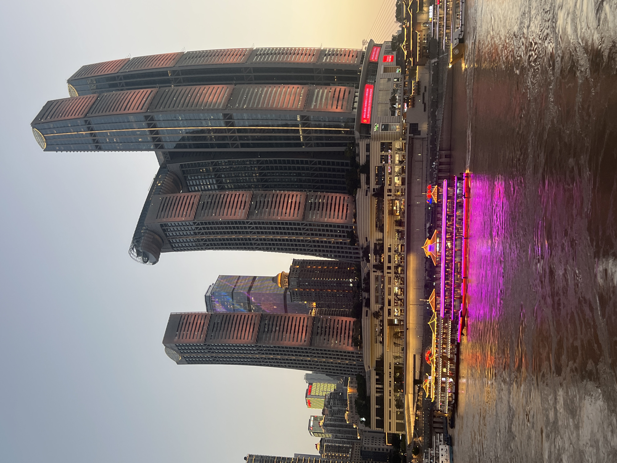The image depicts a twilight scene in Nan'an District, China, likely along a waterfront. Dominating the skyline are several modern skyscrapers, with two particularly tall, twin towers featuring a distinctive reddish-orange hue due to the setting sun reflecting off their facades. The buildings are illuminated, showcasing their architectural details. Below the skyscrapers, a multi-level structure with illuminated windows and balconies stretches along the waterfront, appearing to be a commercial or entertainment complex. In the foreground, a large, ornate tour boat with traditional Chinese architectural elements is brightly lit with pink and yellow lights, casting reflections on the water. Another, smaller white boat is visible further down the river. The water itself is murky brown, with ripples indicating movement. The sky is a soft gradient of pale blue and warm orange, typical of dusk. The overall atmosphere is one of urban vibrancy and nighttime illumination.