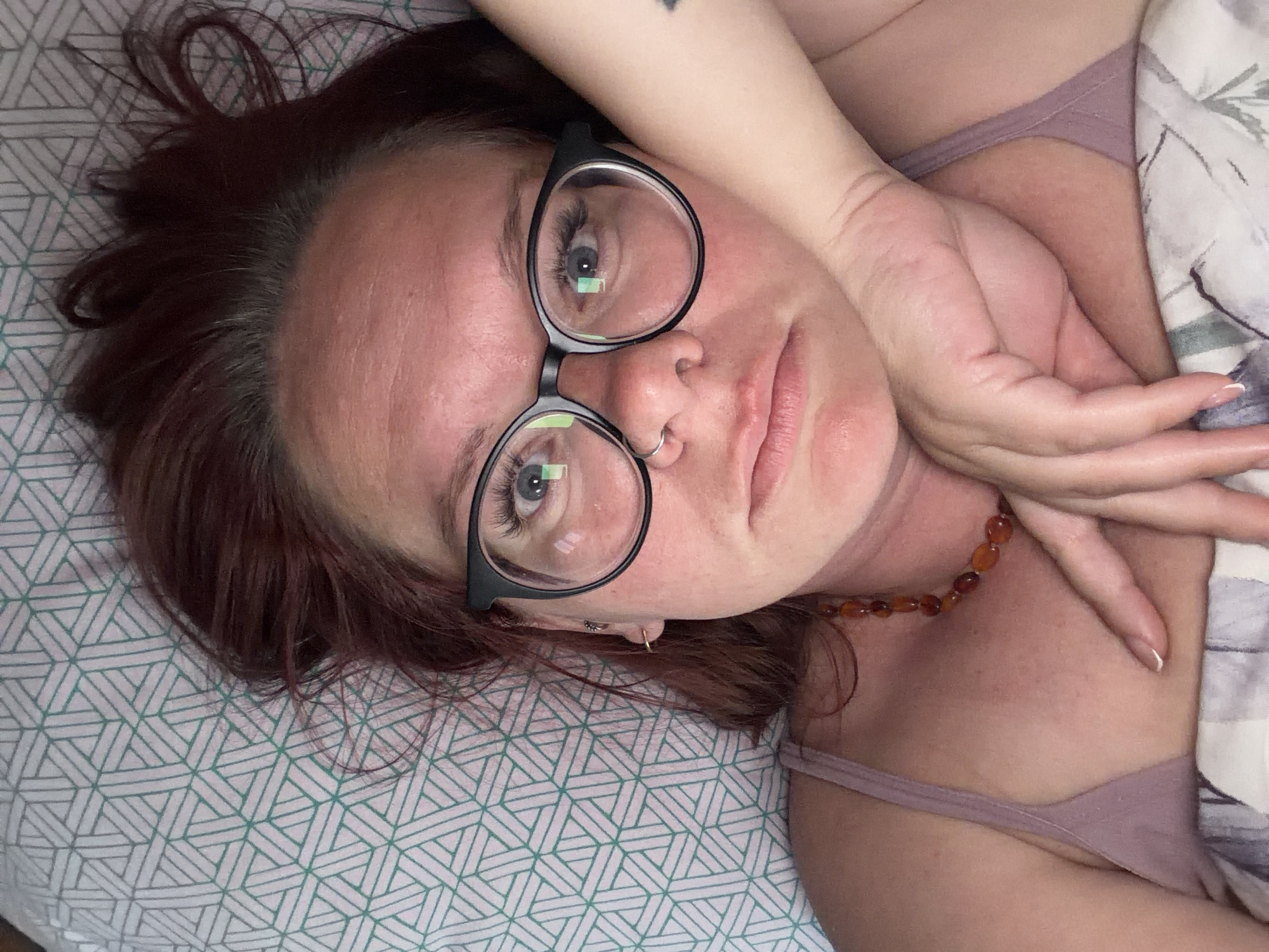 A woman is depicted lying down, looking directly at the viewer, in an indoor setting, likely a bedroom in Chaikovsky, Russia. Her head is tilted to the side, resting on a patterned surface. She has reddish-brown hair, which is fanned out around her head.
She wears large, black-rimmed glasses that frame her light-colored eyes, which are prominent with long eyelashes. A small silver ring pierces her nose, and a delicate gold hoop earring is visible in her left ear. Her complexion is fair, with some natural redness on her forehead and cheeks. Her lips are slightly parted.
One of her arms is raised and bent, with her hand gently resting on her hair or forehead. Her fingernails show a French manicure style. A small, dark, triangular or heart-shaped tattoo is visible on her upper arm. Around her neck, she wears a necklace composed of amber-colored, irregular beads. She appears to be wearing a light-colored, possibly mauve or dusty rose, tank top or bra.
The bedding beneath her head is a light color with a repeating geometric pattern of interconnected triangles and hexagons in a teal or light green hue. To her right, another section of bedding, possibly a duvet or pillow, features a lighter, perhaps white or grey, fabric with a subtle, abstract floral or patterned design. The overall mood conveyed by her expression is relaxed and neutral. There is no visible text in the image.