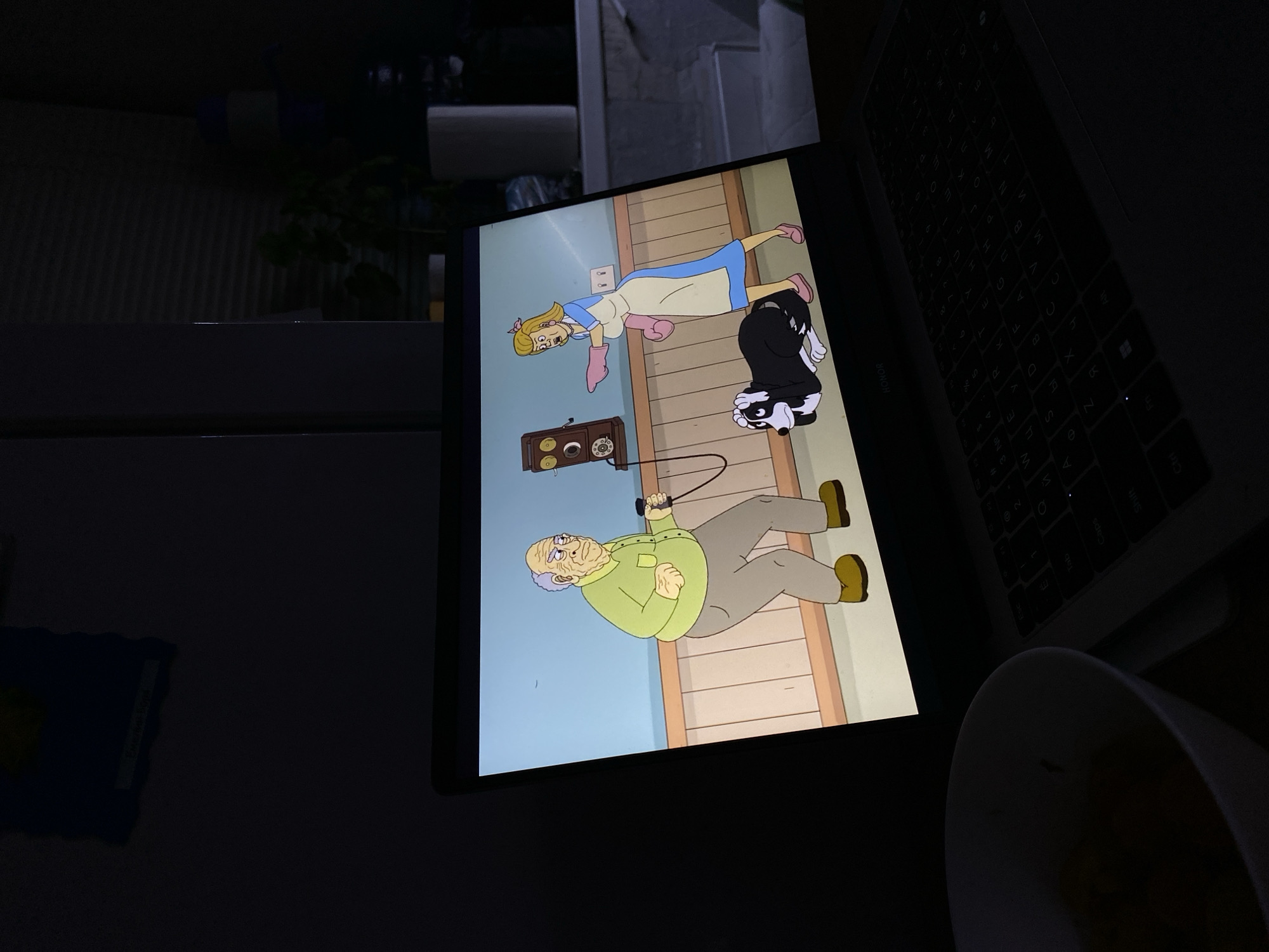 The image shows a screen displaying an animated cartoon. The cartoon features two human characters and a skunk. One character is an elderly man in a green shirt and grey pants, holding a leash. The other is a woman in a blue dress and pink gloves, lying on the floor. A vintage telephone is mounted on the wall.
Next to the screen, a laptop is visible. A cup sits on the surface beside the laptop. The overall lighting in the scene is dim. The location is not specified, and there is no indication of it being in Votkinsk, Russia.