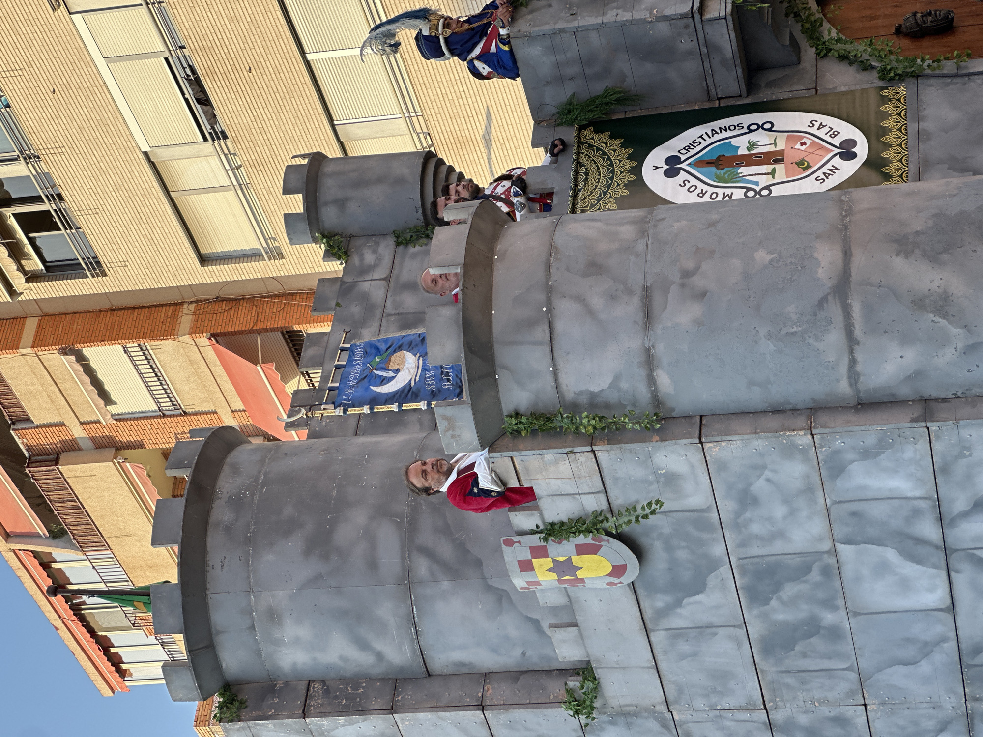 A parade float resembling a castle is depicted in Alicante, Spain, likely during the "Moros y Cristianos" festival. The float is constructed with grey, stone-like panels and features battlements. Several men in historical costumes are visible on the float. One man in the foreground wears a red velvet jacket with white trim and a white ruffled shirt. Another man, to his right, is dressed in a blue and gold military-style uniform with a plumed hat. A banner hangs on the float with the text "MOROS Y CRISTIANOS SAN BLAS" and a crest featuring a tower and palm trees. Green foliage is draped around the castle structure. In the background, modern apartment buildings with balconies and awnings are visible, indicating an urban setting. The sky is clear and blue, suggesting a daytime event.