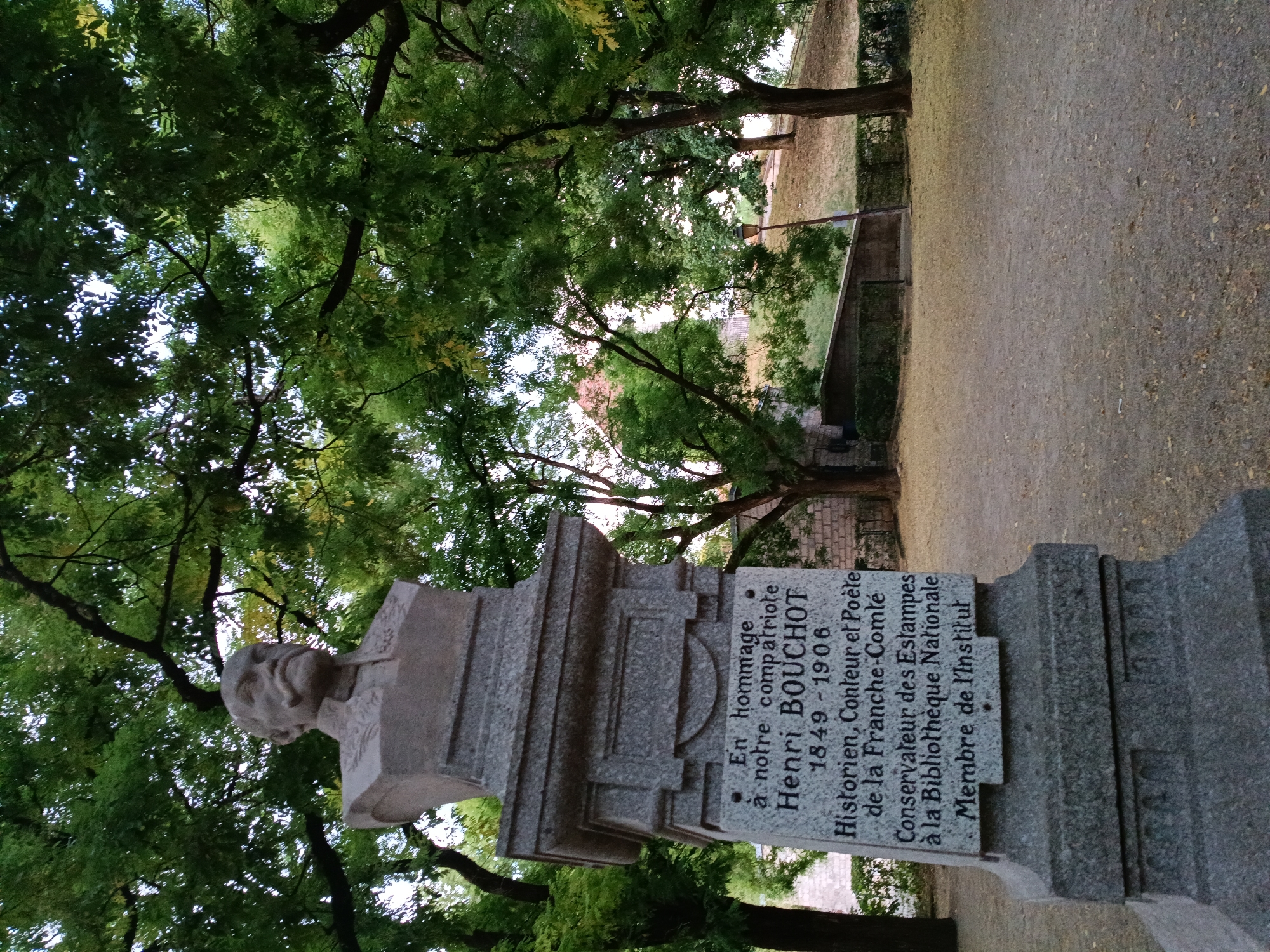 This is a photograph of a stone bust monument dedicated to Henri Bouchot, located in a park in Besançon, France. The bust depicts a man with a mustache, wearing a collared jacket. The base of the monument is inscribed with text detailing his accomplishments as a historian, storyteller, and poet of the Franche-Comté region, as well as his roles as curator of prints at the Bibliothèque Nationale and a member of the Institut. The monument is surrounded by lush green trees, with a path and a lamppost visible in the background. The ground is covered in gravel and fallen leaves.