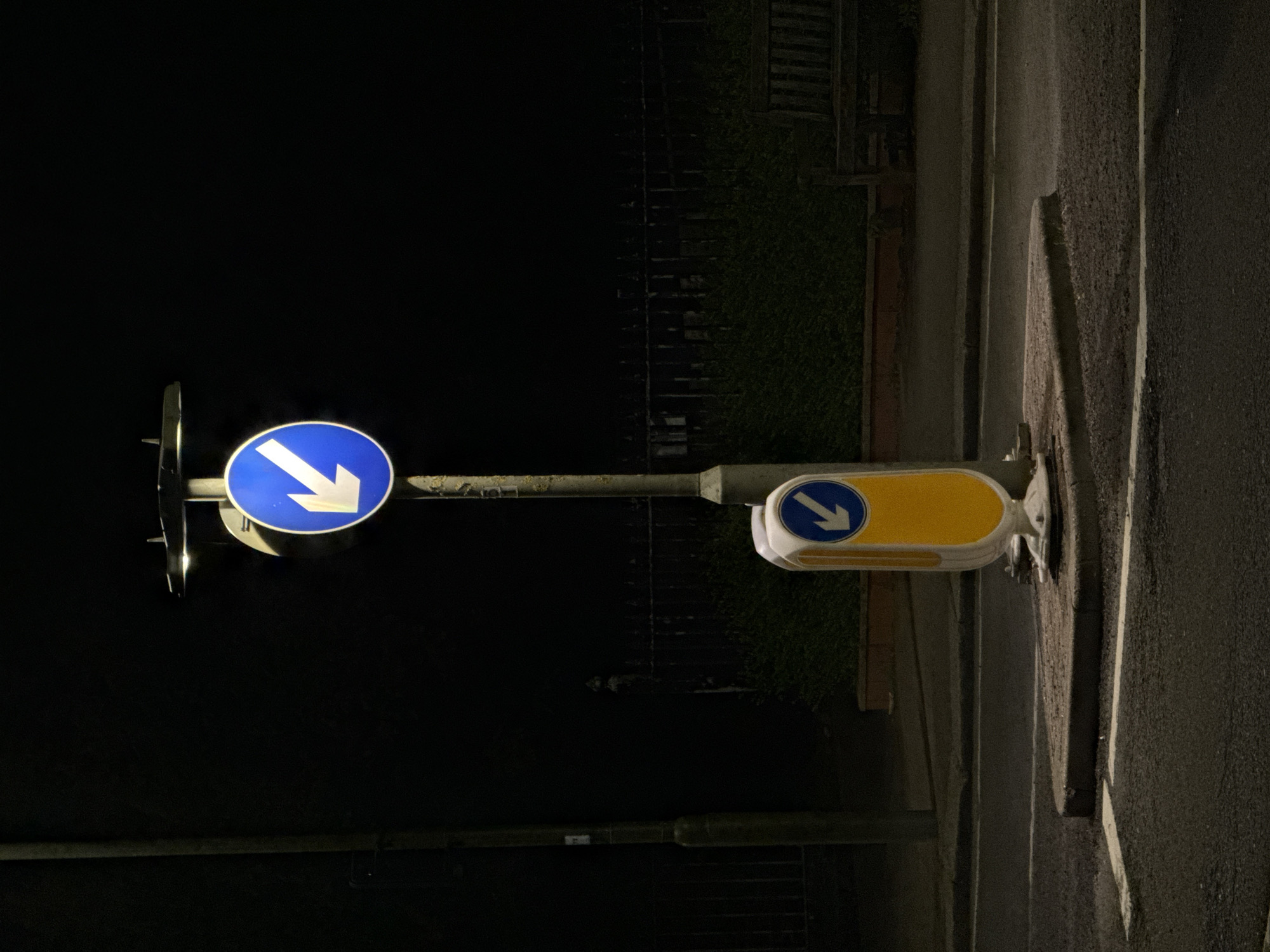 The image captures a nighttime scene on what appears to be a street in London, United Kingdom. The primary subjects are two road signs, both displaying a white arrow pointing diagonally downwards and to the left, indicating a "keep left" or "pass on this side" directive.
The more prominent sign, located towards the left of the frame, is a blue circular sign with a bold white arrow. It is mounted on a simple grey metal pole. To its right, and positioned lower, is a second sign. This one features a yellow, rounded rectangular panel, which encloses a smaller blue circular sign with an identical white arrow pointing diagonally downwards and to the left. This type of yellow-backed sign is commonly used in the UK to mark traffic islands, central refuges, or other obstructions that vehicles must pass on the left.
The scene is dominated by darkness, confirming it is nighttime. The signs themselves appear to be reflective or internally lit, making them clearly visible against the black background. Faint details can be discerned in the background, including what looks like dark foliage, possibly a hedge, and some indistinct structures like a fence or wall, all shrouded in shadow. To the far right, a dark asphalt road surface is visible, bordered by a curb or pavement edge. There are no people, vehicles, or discernible weather conditions. The street appears quiet and devoid of activity. No visible text is present on the signs, only symbolic arrows.