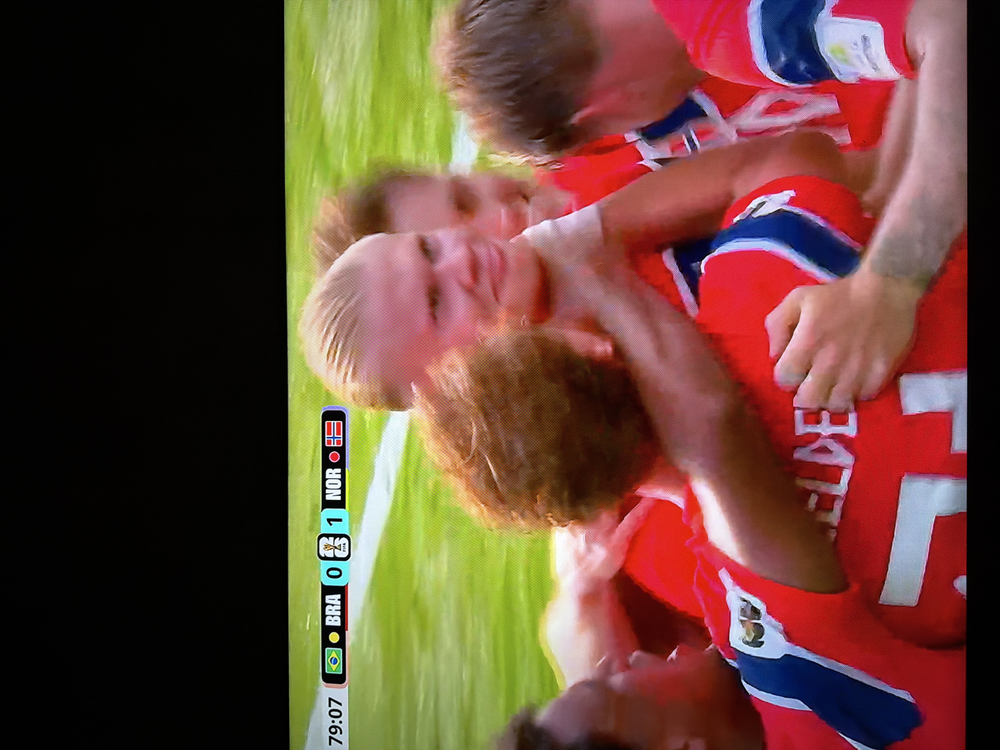 The image shows a soccer match scoreboard with Brazil (BRA) at 0 and Norway (NOR) at 1, at the 79:07 minute mark. Several soccer players in red jerseys are celebrating a goal, with one player, seemingly Erling Haaland, in the foreground with his head turned towards the camera and a smile. The background consists of a bright green soccer field. The location is not specified in the image or crops.