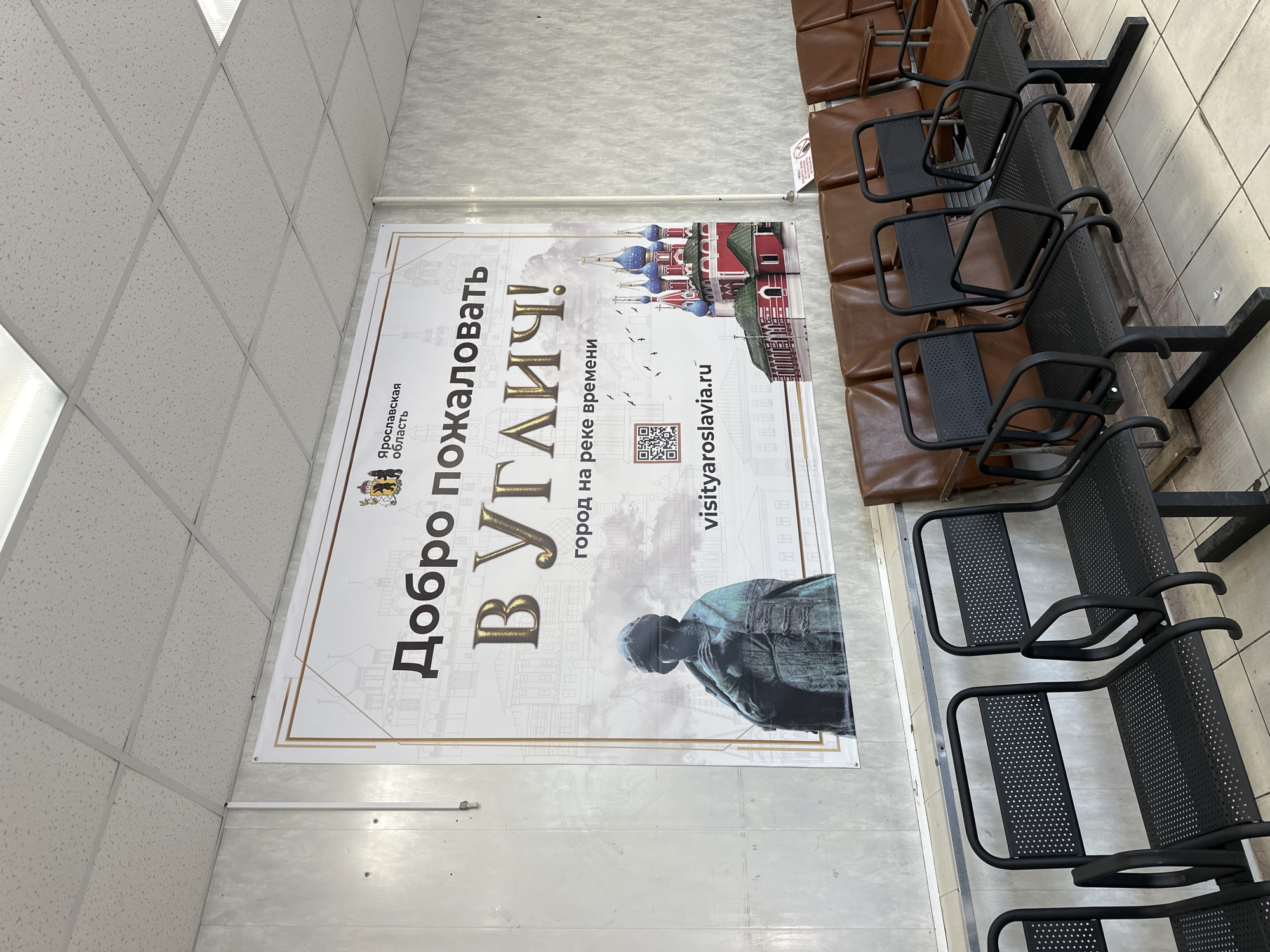 A large banner hangs on a wall, featuring an image of the city of Uglich, Russia, with the text "Добро пожаловать В УГЛИЧ!" (Welcome to Uglich!) and "город на реке времени" (city on the river of time). The banner also displays the website "visityaroslavia.ru" and a QR code. In the foreground, several rows of chairs are visible. The chairs in the front are black, metal chairs with perforated seats and backs, and curved armrests. Behind these are rows of brown leather chairs. The environment appears to be an indoor waiting area.