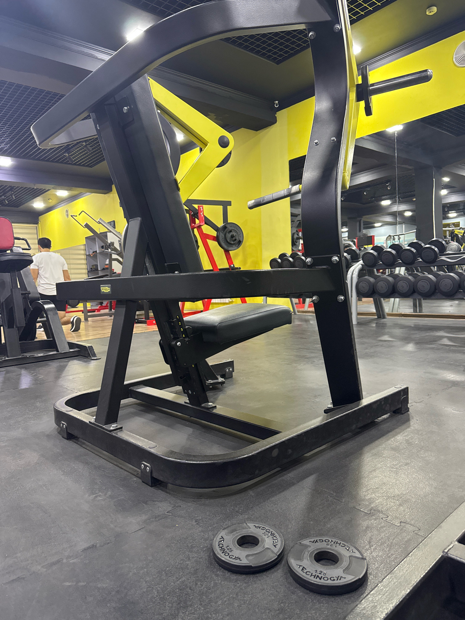 In a well-lit, modern gym in Tashkent, Uzbekistan, a black Technogym weight machine occupies the foreground. The machine features a padded black seat and a robust black frame, with yellow accents visible on its structure and in the background. The floor is covered with grey interlocking mats. Two small, circular weight plates, each labeled "1.25 TECHNOGYM," are placed on the mat in the lower right quadrant of the image. In the background, a person wearing a white t-shirt and black shorts is visible, seemingly engaged in a workout. Additional gym equipment, including a rack of dumbbells and other weight machines, are present in the blurred background, contributing to the overall ambiance of a functional fitness facility. The walls of the gym are painted a bright yellow, and a mirrored wall is visible in the distance.