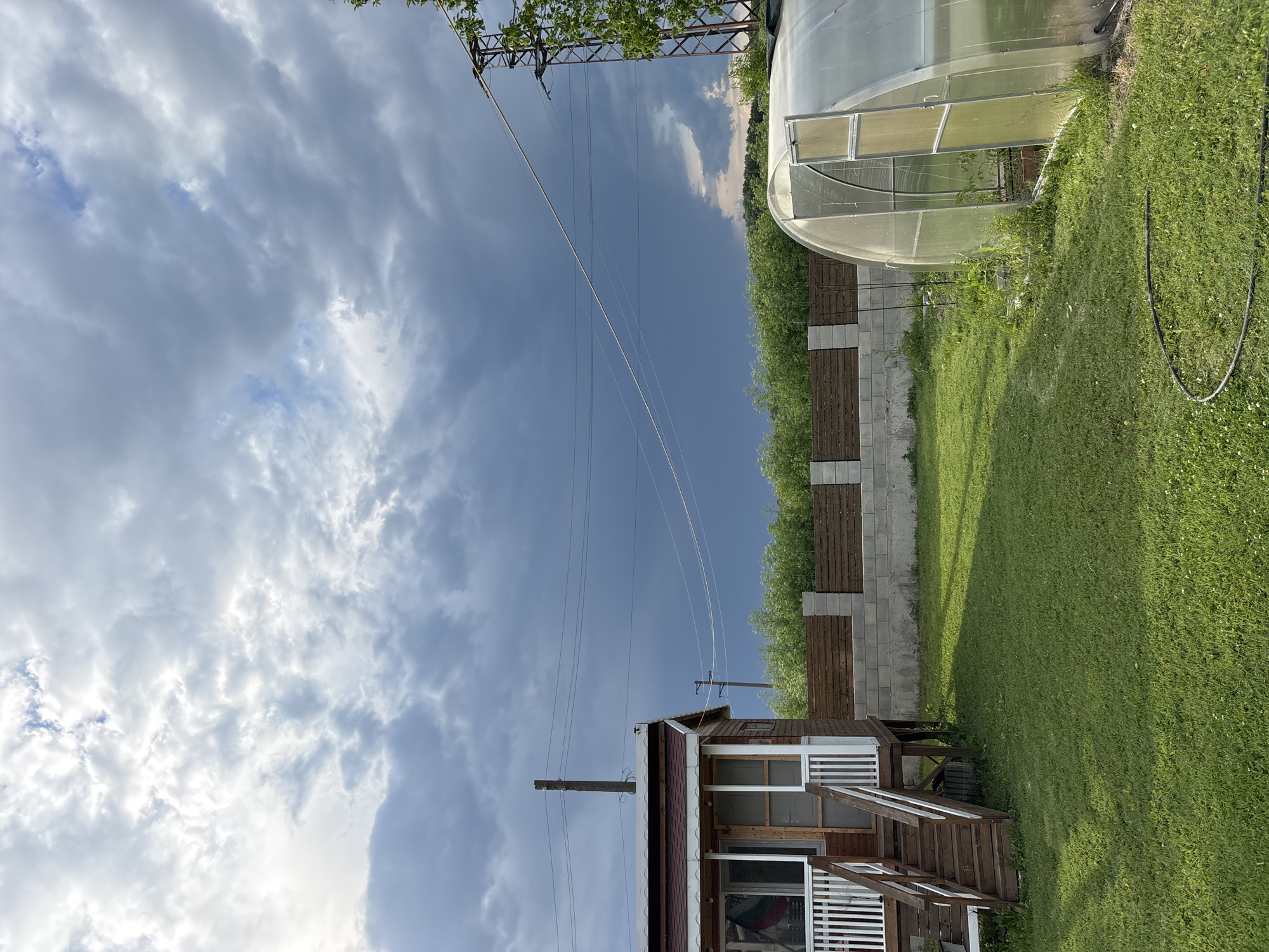 The image presents an outdoor scene, potentially located in a rural or suburban setting near Pchelnik, Bulgaria. The sky is filled with a mix of grey and white clouds, suggesting varying weather conditions. Power lines stretch across the upper portion of the frame. The right side of the image shows a green lawn and a greenhouse structure. A fence, made of concrete blocks and wooden planks, borders the lawn. In the lower part of the frame, a small wooden house with white trim is visible. An open door provides a glimpse into the interior.