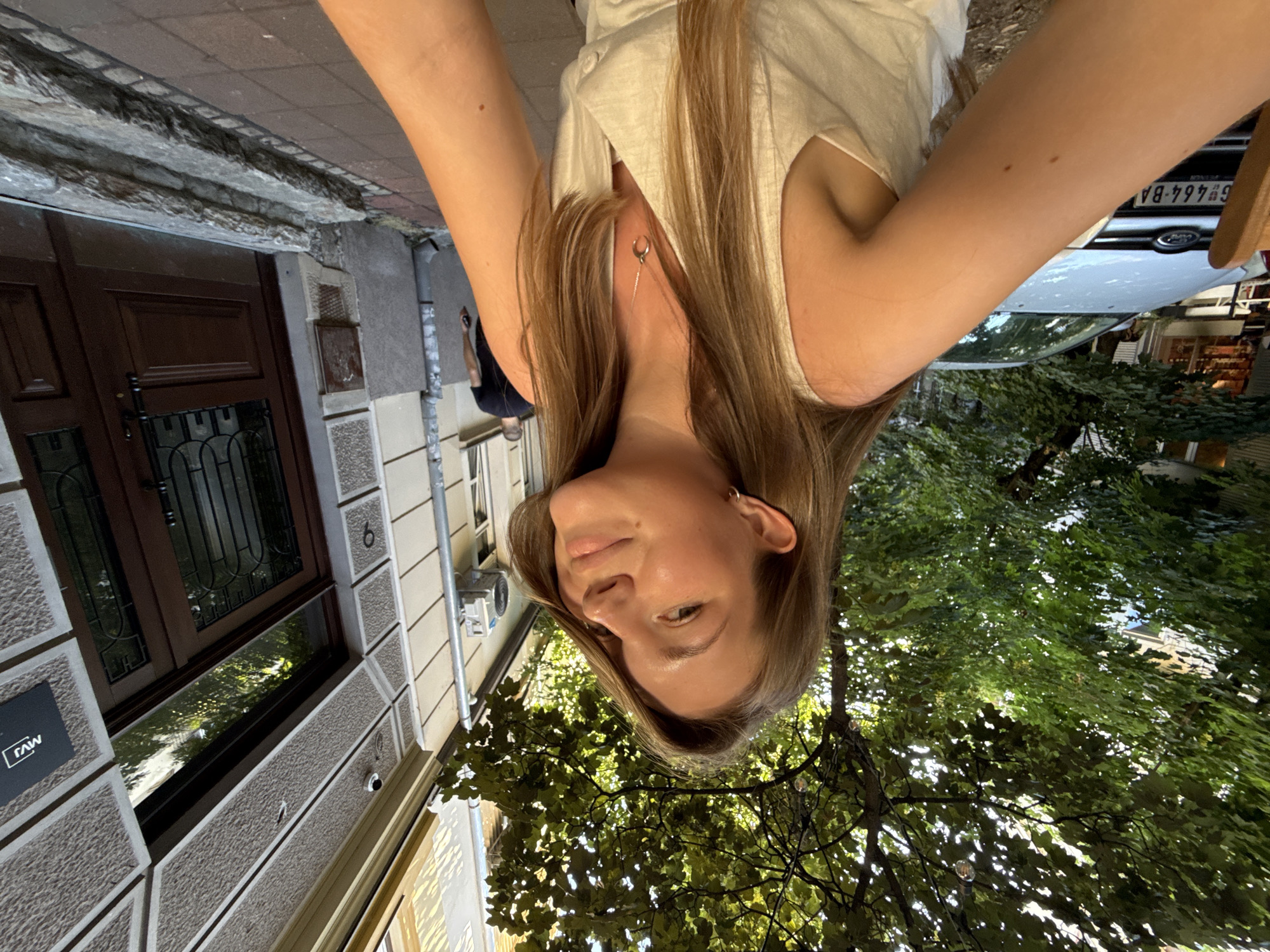 The image captures a young woman in the foreground, her face angled towards the camera with a relaxed smile. She has long, light brown hair and is wearing a white sleeveless top, a delicate silver necklace with a crescent moon pendant visible at her collarbone. She is outdoors, with a tree providing dappled shade over the scene, suggesting an outdoor seating area of a cafe or restaurant. Behind her, to the left, a white car is parked, its license plate reading "G 464-BA". The background features a building with a textured facade, dark brown double doors, and stone steps leading up to the entrance. A sign with "MVJ" in white letters on a black background is visible on the building's facade. Further in the background, a man in a black t-shirt walks away from the camera. The presence of the car and the architecture of the building indicate an urban setting, likely a street or a courtyard in Belgrade, Serbia.