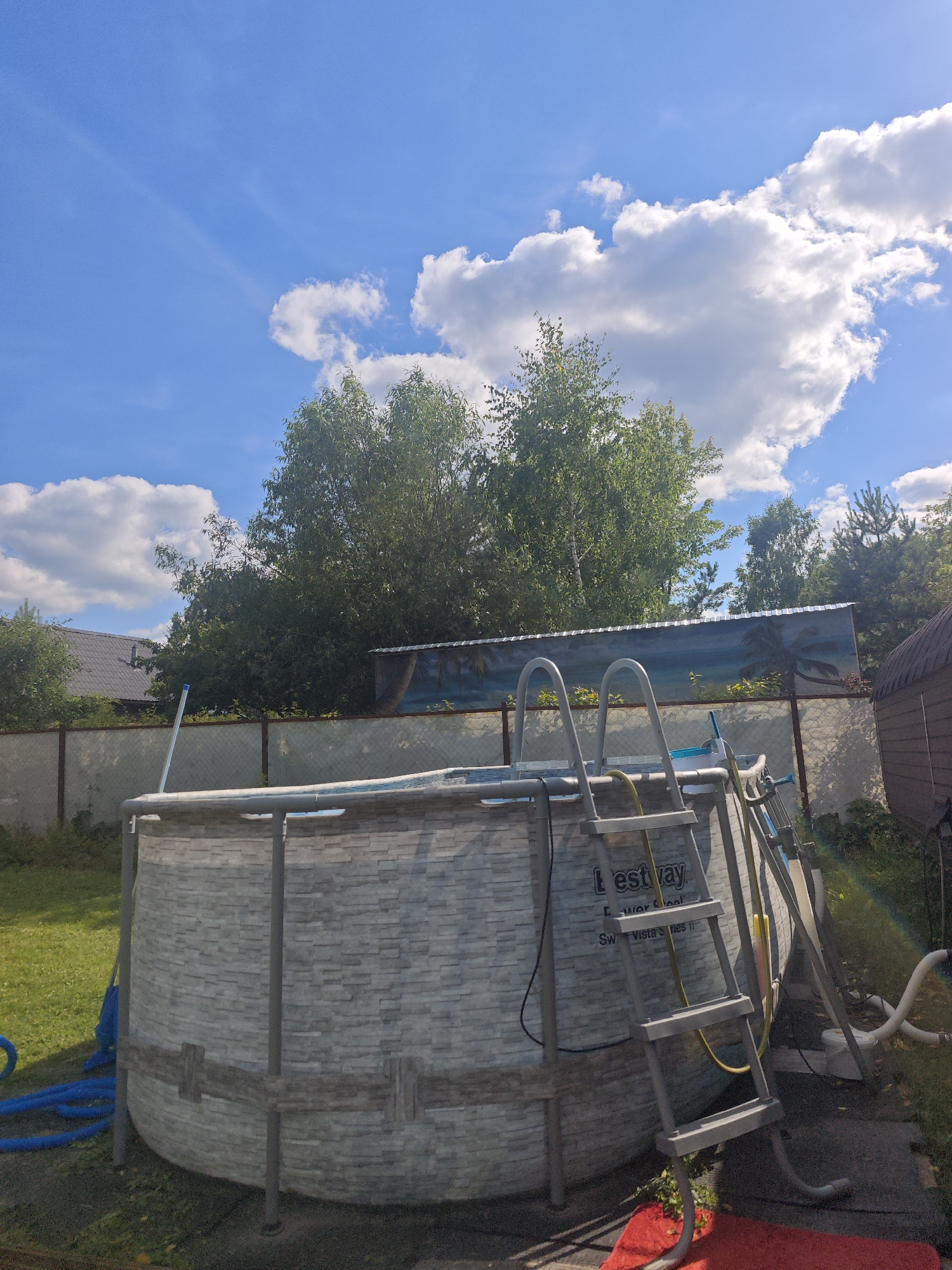 The image captures an outdoor scene on a sunny day with a bright blue sky and scattered white clouds, suggesting it's midday. The setting is a backyard in Khlevino, Russia, characterized by a well-maintained lawn and lush green trees forming a backdrop.
The central object is a large, round, above-ground swimming pool with a stone-patterned exterior and a metallic frame. A silver ladder with gray steps is attached to the pool, providing access. There are also blue hoses and white piping connected to the pool's filtration system, indicating the pool is functional. A red mat lies on the ground near the ladder. In the background, a chain-link fence encloses the yard, and behind it, a residential building with a dark roof is visible. A weathered wooden shed or structure stands to the right of the pool. A decorative banner depicting palm trees and a beach scene is attached to the fence.
No people are visible in the image. The overall mood is peaceful and suggests a warm, pleasant day, ideal for swimming. The visible text on the pool reads "Bestway" and "Power Steel."