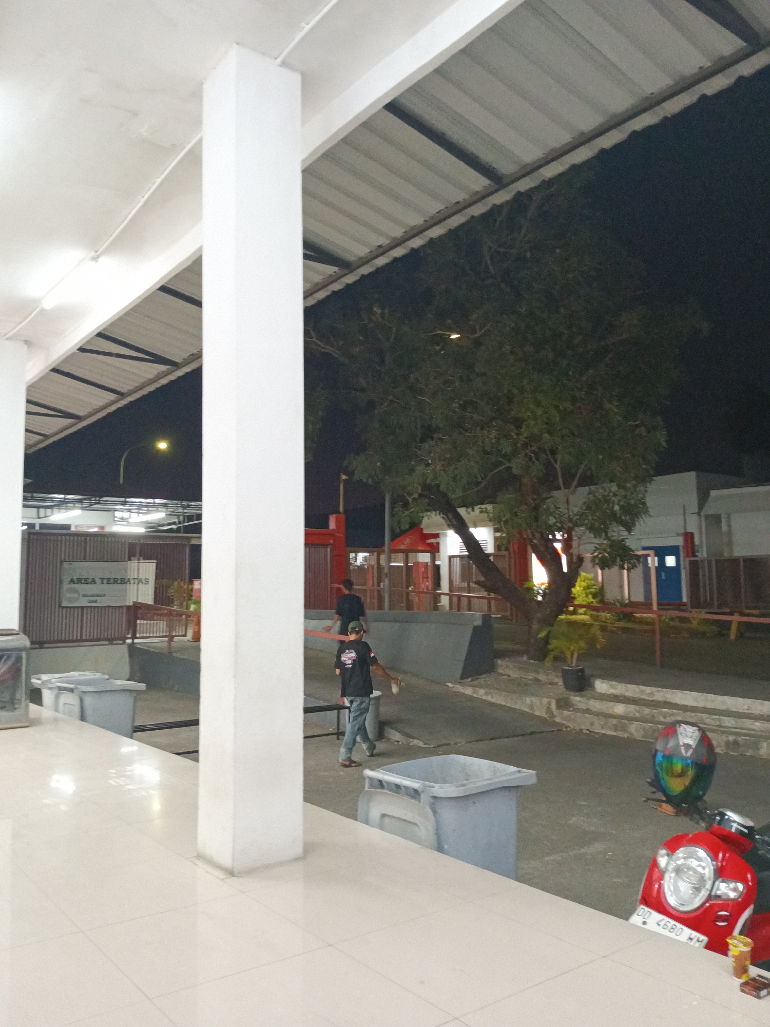 The image captures a dimly lit outdoor area at night, possibly within a facility or compound in Makassar, Indonesia. In the foreground, a large white pillar dominates the left side of the frame, with a tiled floor extending outwards. Beyond the pillar, a sign reads "AREA TERBATAS" (Restricted Area) on a corrugated metal fence. Two large grey trash bins are visible in the middle ground.
To the right, a person wearing a black shirt and blue jeans is walking away from the camera, pushing a cart or broom. Further back, a large tree with dense foliage stands prominently, illuminated by an unseen light source. Behind the tree, a red motorcycle is parked, featuring a colorful helmet resting on its seat and a visible license plate "DD 4680 WH". The background consists of buildings with visible windows and doors, some with red accents. The overall impression is of a service or utility area after dark.