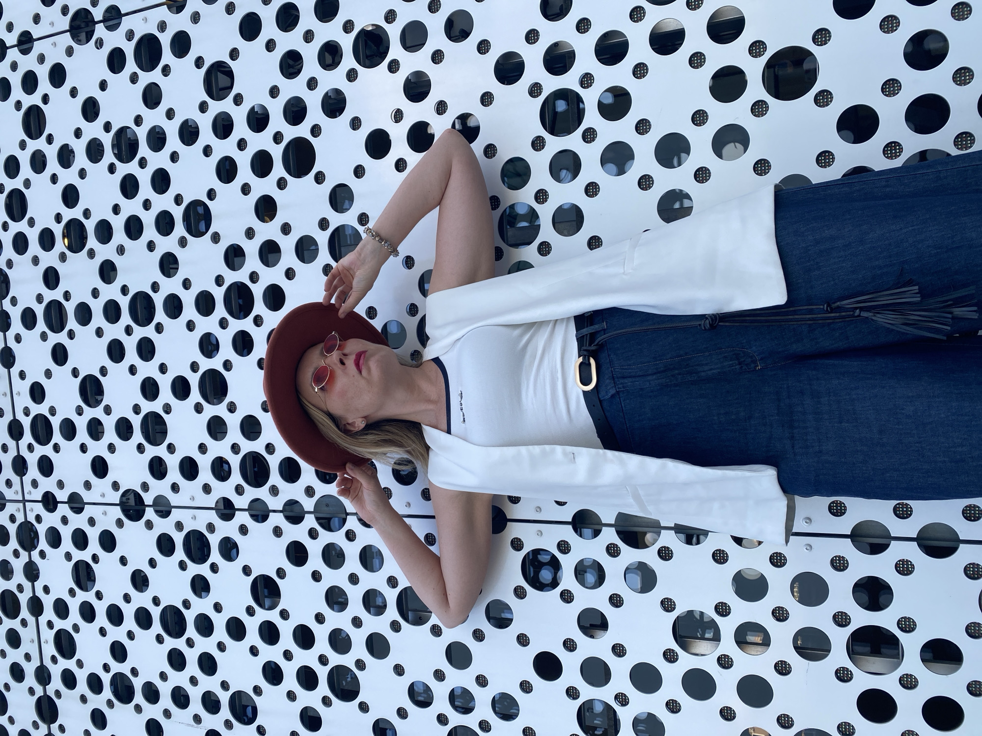 A woman is lying on a white surface with black circular cutouts. The surface appears to be part of a building facade. The woman is wearing a red hat, sunglasses, a white tank top, a white sleeveless jacket, dark denim jeans with a black belt. She is positioned diagonally across the frame, with her head towards the upper left corner. Her right hand is raised and slightly touching her hat. The background shows elements of the city Yekaterinburg, Russia, visible through the circular cutouts.