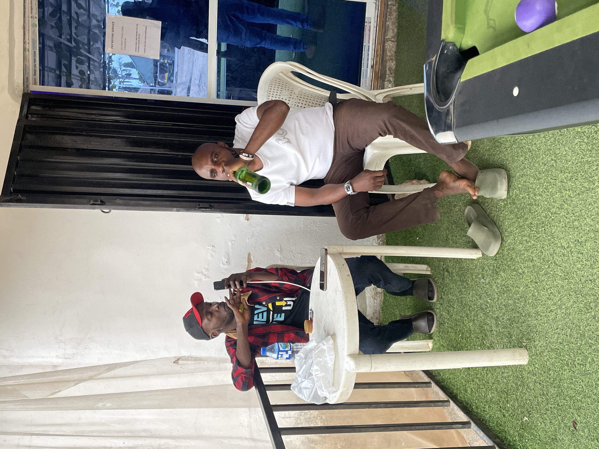 Two men are sitting outdoors, possibly in a backyard or patio area. The man at the top is drinking from a green glass bottle, wearing a white shirt, brown pants, a watch, and green sandals. He is seated in a white plastic chair, and a partially visible pool table with a purple ball is beside him. The man at the bottom is wearing a red and black plaid shirt over a black t-shirt, black pants, a red and black baseball cap, and dark sandals. He is holding a phone and something in his other hand, possibly smoking. He is also seated in a white plastic chair, and there is a small table with a water bottle and a plastic bag in front of him. The background includes a white wall, a black metal structure, artificial green turf, and a window with a sign that reads "PAY BEFORE SERVICE. NO CREDIT TODAY COME TOMORROW". The location is possibly in Barakin Akawo, Nigeria.