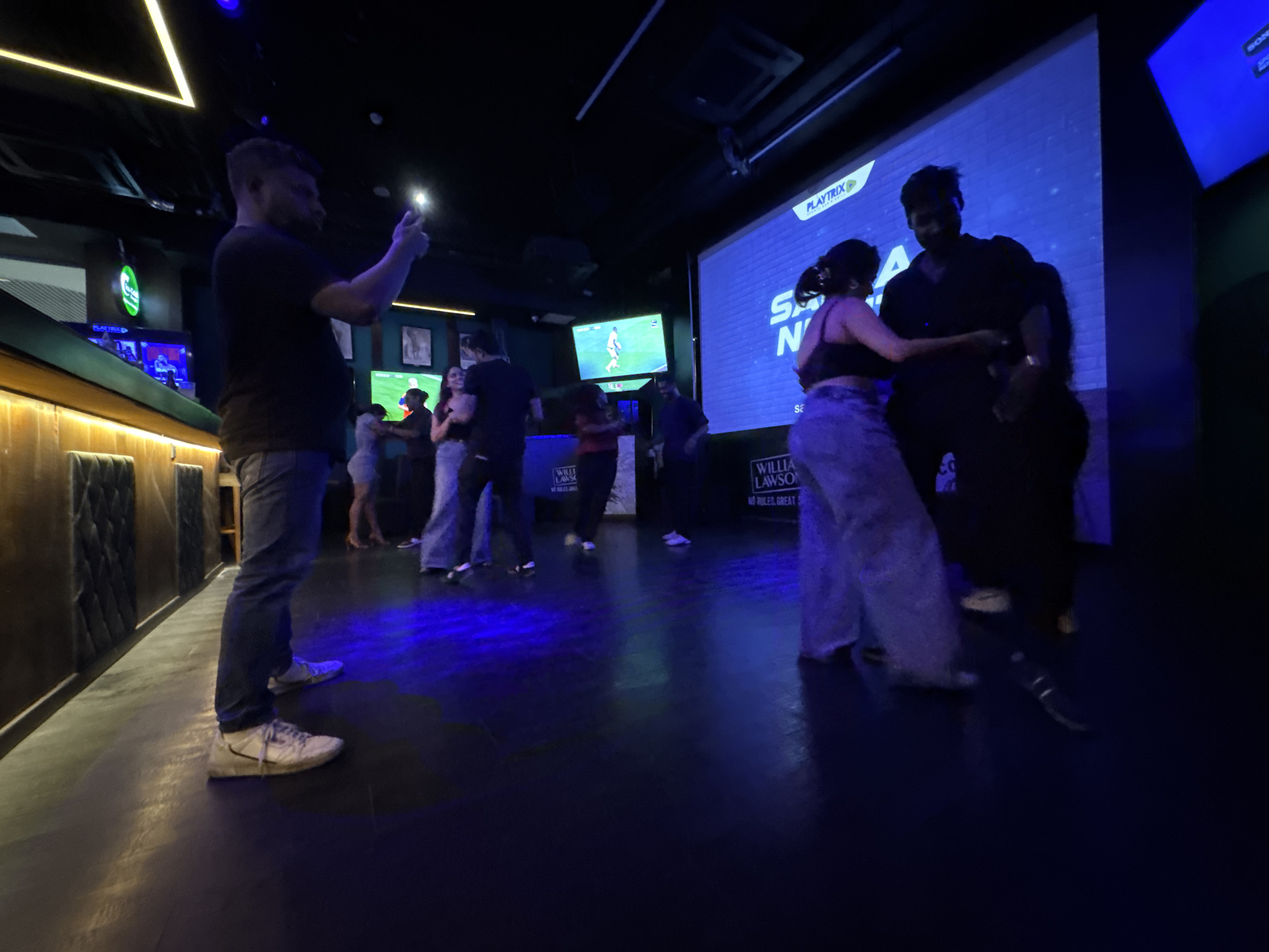 This image captures a vibrant night scene inside a bar or club in Colombo, Sri Lanka. The setting is dimly lit with predominantly blue and white artificial lighting, creating an energetic atmosphere. The floor is dark, suggesting a dance area.
In the left foreground, a man wearing a black t-shirt, blue jeans, and white sneakers stands with his back partially to the viewer, holding up a smartphone with its light on, possibly recording or taking a photo. To the right, a couple is engaged in dancing; the woman wears a dark crop top and light-colored wide-leg pants, and the man is in dark attire. Several other individuals are visible in the background, also socializing and dancing, some in motion blur.
Notable details include a long bar or counter with dark wooden paneling and upholstered seating on the left. Multiple large screens are prominent in the background. The largest screen displays the "PLAYTRIX" logo, with white text that appears to read "SALSA NIGH" (likely "Salsa Night"), and "WILLIAM LAWSON'S" followed by "NO RULES. GREAT S" (suggesting a sponsor like William Lawson's Scotch). Another smaller screen in the mid-background shows what looks like a football match. A bright, yellow-white triangular light fixture is visible on the ceiling on the left. The scene evokes a lively, social event or party.