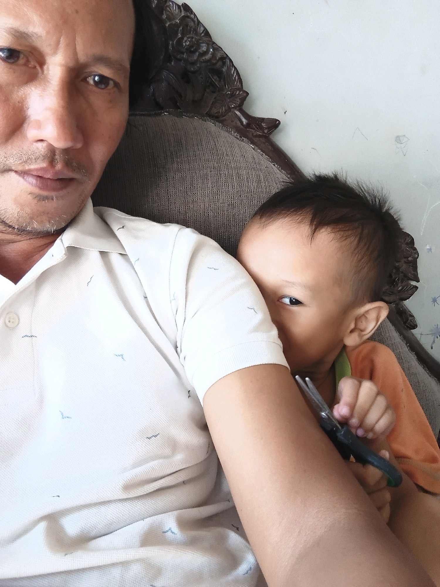 The image shows a close-up of a man and a child in an indoor setting. The man, partially visible on the left, has dark hair and a mustache, and is wearing a light-colored collared shirt with a subtle pattern. The child, on the right, is peeking out from behind the man's arm, with dark hair styled upwards. The child is wearing an orange shirt and holding a pair of scissors. They are both seated on a sofa with a textured fabric. In the background, a wall is visible, with some faint marks or drawings on it. The overall impression is a candid moment, possibly in a home environment. The location is identified as Cikal, Indonesia.