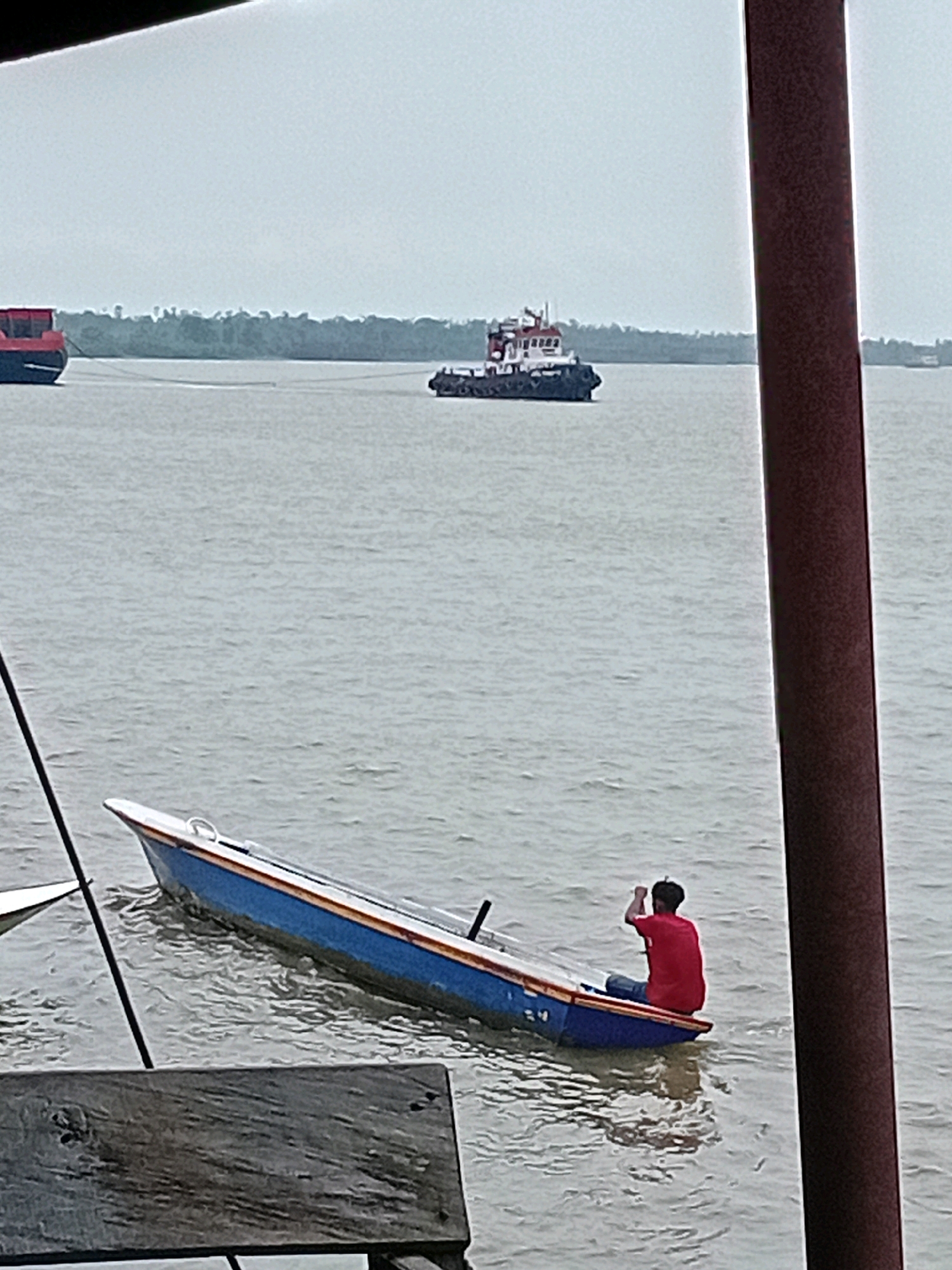 A person in a red shirt and blue jeans is seated in a small, partially submerged blue and orange boat on a body of water, likely the river near Sungai Meriam, Indonesia. The person is facing away from the camera and appears to be gesturing with their right arm. In the background, a tugboat is visible in the distance, and a large red and blue cargo ship is partially visible on the left. The water is choppy, and the sky is overcast. A dark, reddish-brown pole stands prominently in the foreground on the right side of the image, and a wooden surface is visible in the lower left corner, obscuring part of the scene. The overall atmosphere suggests a cloudy or hazy day.