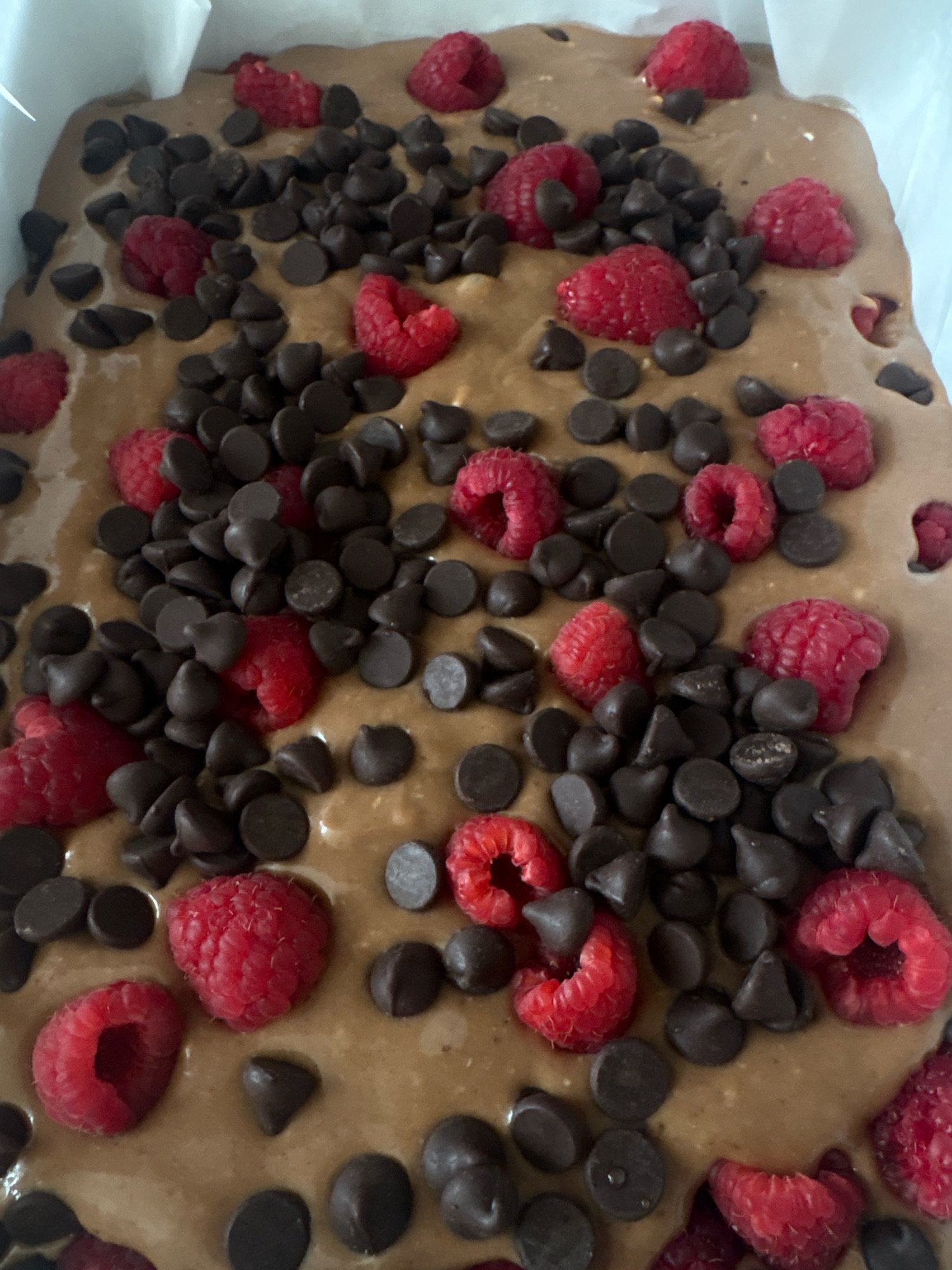 The image shows a close-up, overhead view of a baked good, possibly brownies or a cake, in a white baking dish. The batter is a light brown color, and it is studded with numerous dark chocolate chips and whole raspberries. The raspberries are bright red and appear fresh, some are whole, and others are cut in half. The chocolate chips are dark brown and have a typical semi-sweet chocolate chip shape. The baked good appears to be freshly made and is presented in a way that suggests it is ready to be served or is in the process of being prepared for consumption. There are no people visible in the image. The environment is not discernible beyond the baking dish itself. The location of this scene in Mexico City, Mexico, cannot be determined from the visual information provided.