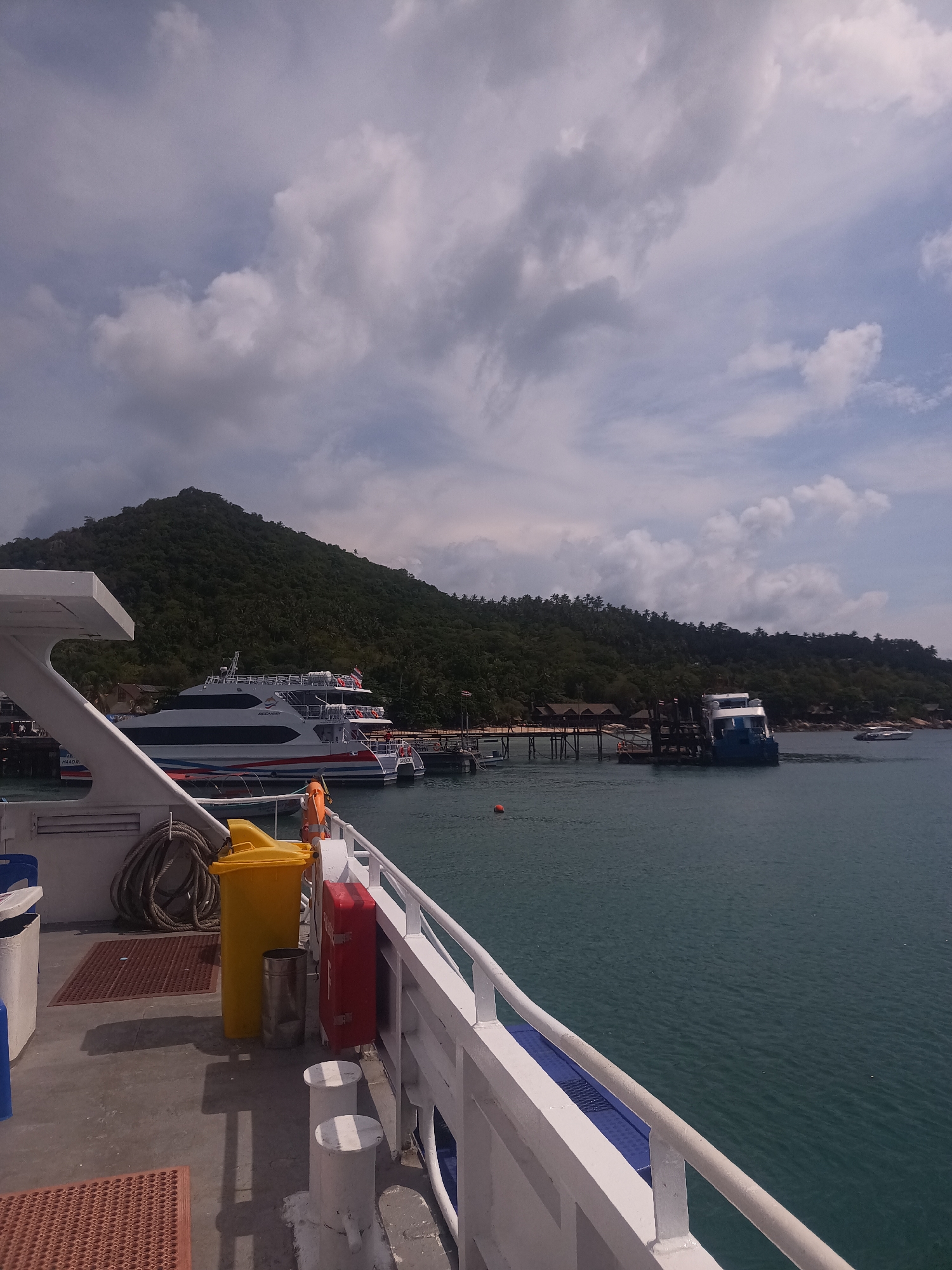 The image depicts a view from the deck of a boat or ferry in Tambon Ko Tao, Thailand. In the foreground, portions of the deck are visible, including a yellow trash bin, a red fire hose cabinet, and various mooring posts. A coil of rope is also visible.
The midground features a body of water, most likely the sea, with other boats and ferry vessels docked in the distance. A small pier or landing is connected to the shore.
In the background, a lush, green, mountainous island or peninsula rises prominently, partially obscured by a sky filled with scattered white and gray clouds. The overall impression is one of a coastal scene on a partly cloudy day.