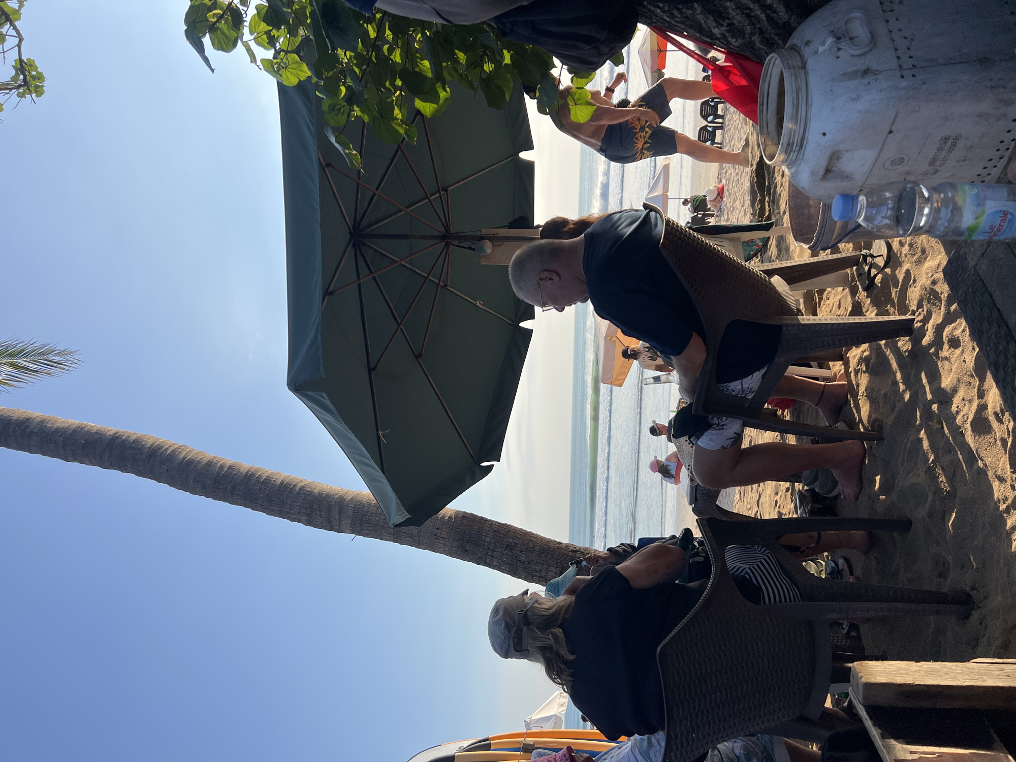 The image depicts a beach scene in Kuta, Indonesia, during daylight hours. In the foreground, two individuals are seated at a table on the sand. One person, wearing a grey cap and black t-shirt, faces away from the camera. The other, wearing glasses and a dark t-shirt, is also facing away and appears to be looking towards the ocean. A large green umbrella provides shade over their seating area.
In the middle ground, the ocean with gentle waves is visible, and several other beachgoers are present, some seated and others walking. A man in floral patterned shorts is walking in the background.
The environment is characterized by sand, a tall palm tree, and lush green foliage. The sky is clear and blue. Objects like chairs, a table, surfboards (partially visible on the left), a red bag, and a large white container with a blue-capped water bottle are present. The overall atmosphere suggests a relaxed beachside dining or lounging experience.