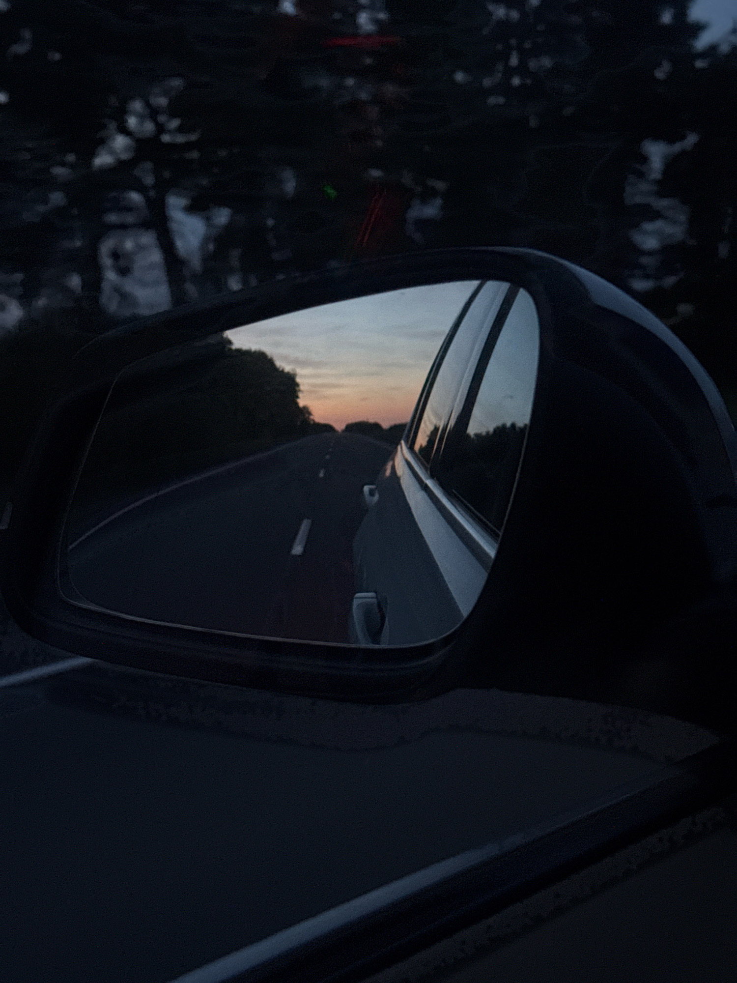 The image captures a view through a car's side mirror, reflecting a road at dusk. The mirror shows a dark asphalt road with white lane markings, curving gently to the right. Trees and foliage line the sides of the road, silhouetted against a soft, twilight sky with hints of orange and blue. The car itself is visible in the foreground, its dark, sleek exterior reflecting the surrounding environment. The overall impression is one of a quiet, solitary journey on a country road as day transitions into night. The location, Васькавіцкі сельскі Савет, Belarus, suggests a rural setting, which aligns with the imagery of a road bordered by trees and open sky.