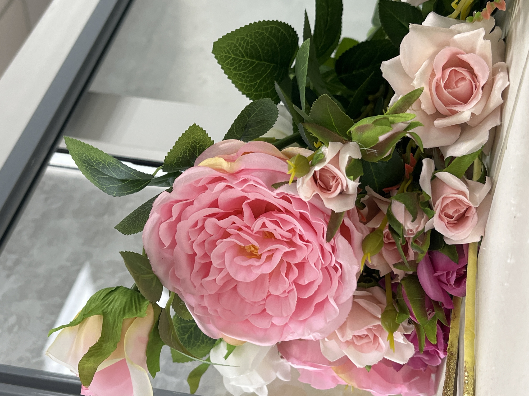 A close-up shot captures a bouquet of artificial flowers, primarily roses, arranged in various shades of pink, white, and deep magenta. The blooms are densely packed, with some fully open and others in bud. Lush green artificial leaves intersperse the flowers, adding a touch of contrast. The bouquet rests on a light-colored surface, possibly a shelf or table. In the background, a window with a grey frame is visible, reflecting light and the surrounding environment. The scene is indoors, and the overall lighting suggests a moderately lit room. There are no people or animals present in the image, and no specific landmarks or contextual clues suggest a precise location within Ridder, Kazakhstan. The image focuses solely on the floral arrangement and its immediate surroundings.