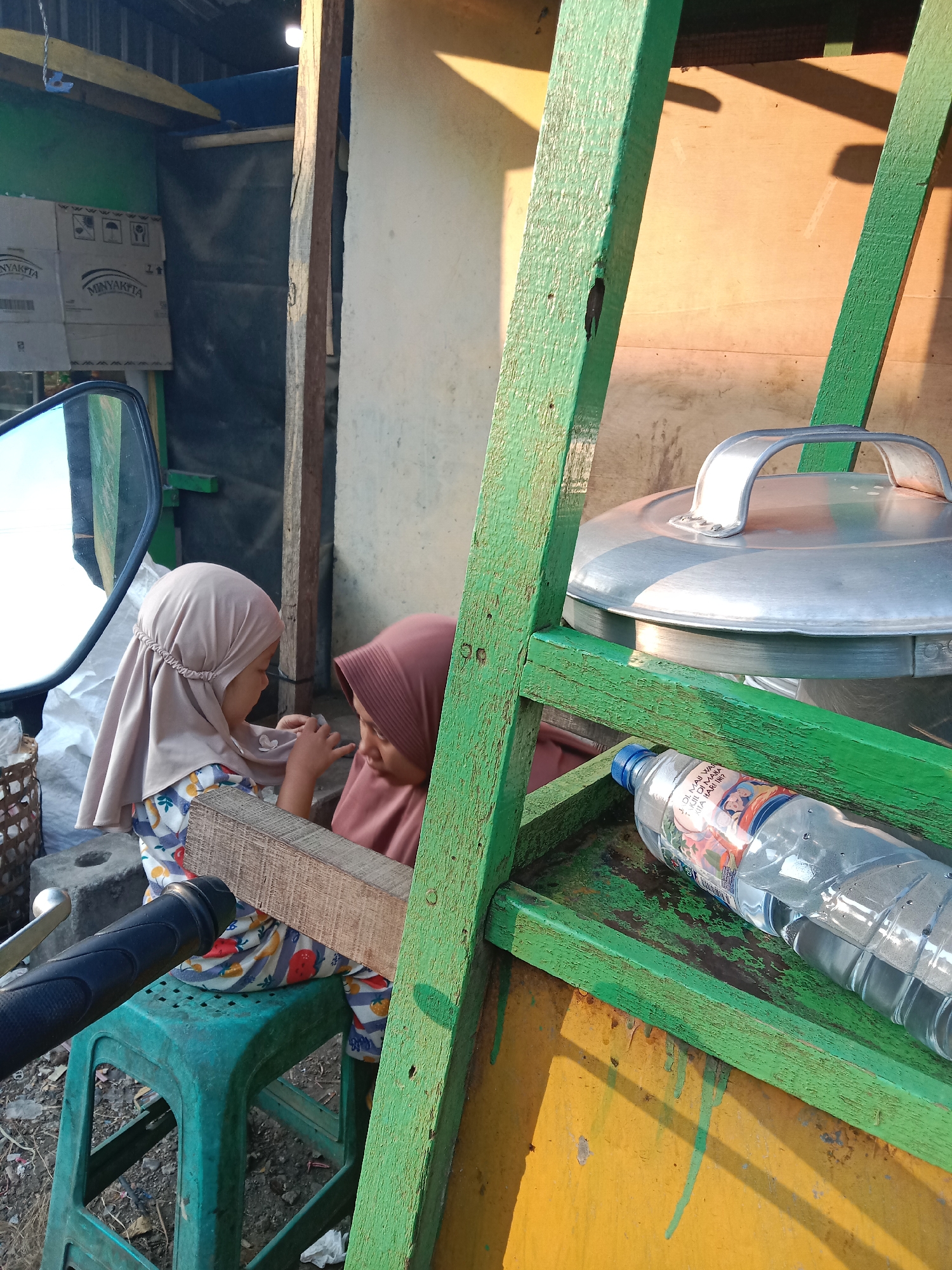 In Sukomulyo, Indonesia, a sun-drenched outdoor scene features two young girls wearing hijabs. One girl, in a light brown hijab and a patterned shirt, sits on a green plastic stool. The other girl, in a darker pink hijab, is partially visible to her right. They appear to be interacting with a wooden plank positioned horizontally between them.
To the right of the girls, a large, silver lidded pot rests on a green wooden shelf. Below this shelf, a clear plastic bottle of water, with a label featuring children's images, lies on its side. The environment appears to be an outdoor market or roadside stall, with a corrugated metal roof structure visible in the background. Cardboard boxes labeled "MINYAKITA" are stacked against a wall. A side mirror from what appears to be a motorcycle is reflected on the left side of the frame. The ground is dirt and debris. The overall lighting suggests a bright, sunny day.