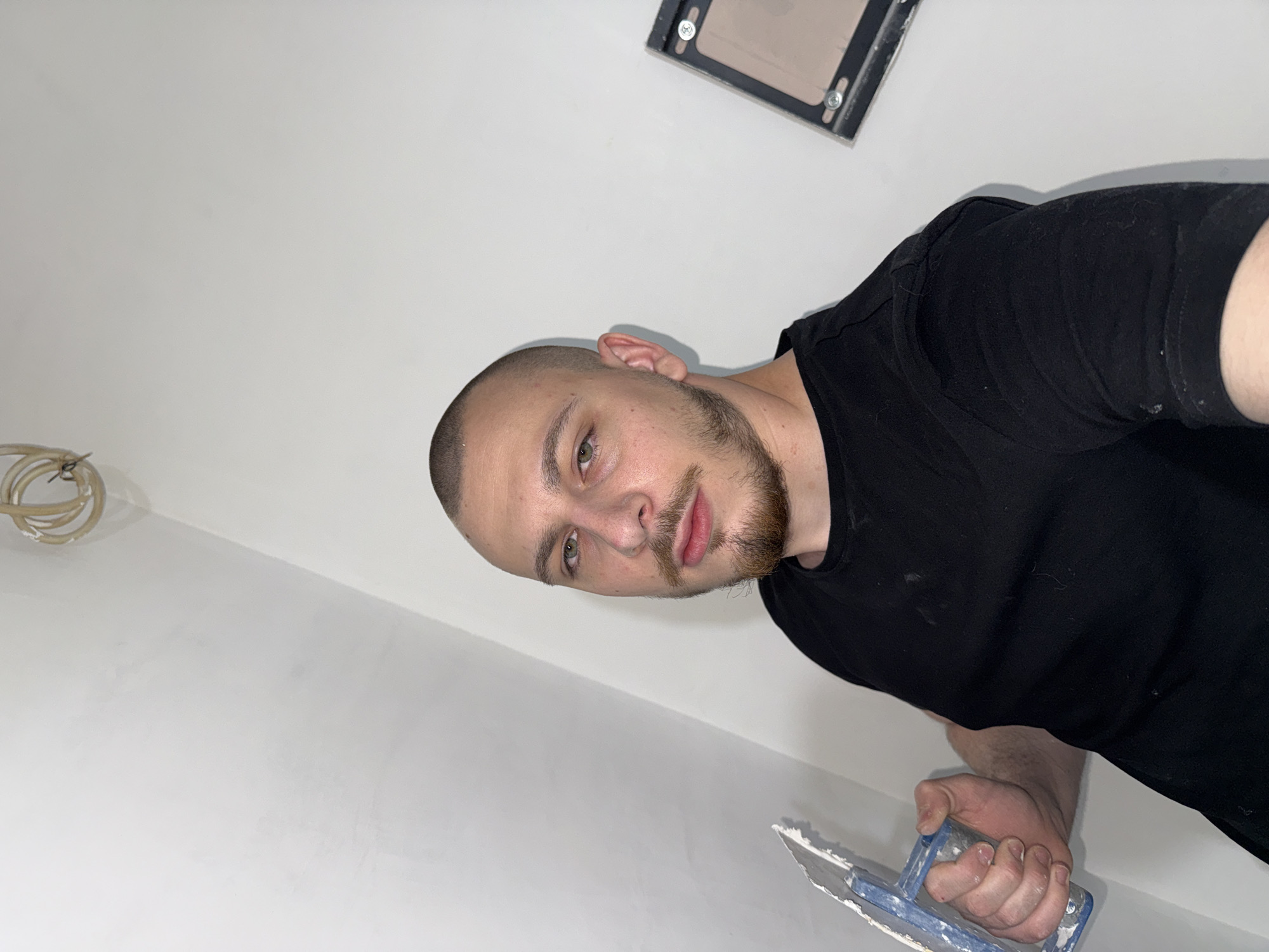 A man with a shaved head and a beard is standing in a room with white walls. He is holding a blue-handled trowel in his right hand. There is exposed wiring on the wall to his left. The man is wearing a black t-shirt. There is a rectangular electrical component mounted on the wall in the top center of the frame. This image does not contain information to determine a specific location within the city of Stara Zagora, Bulgaria.