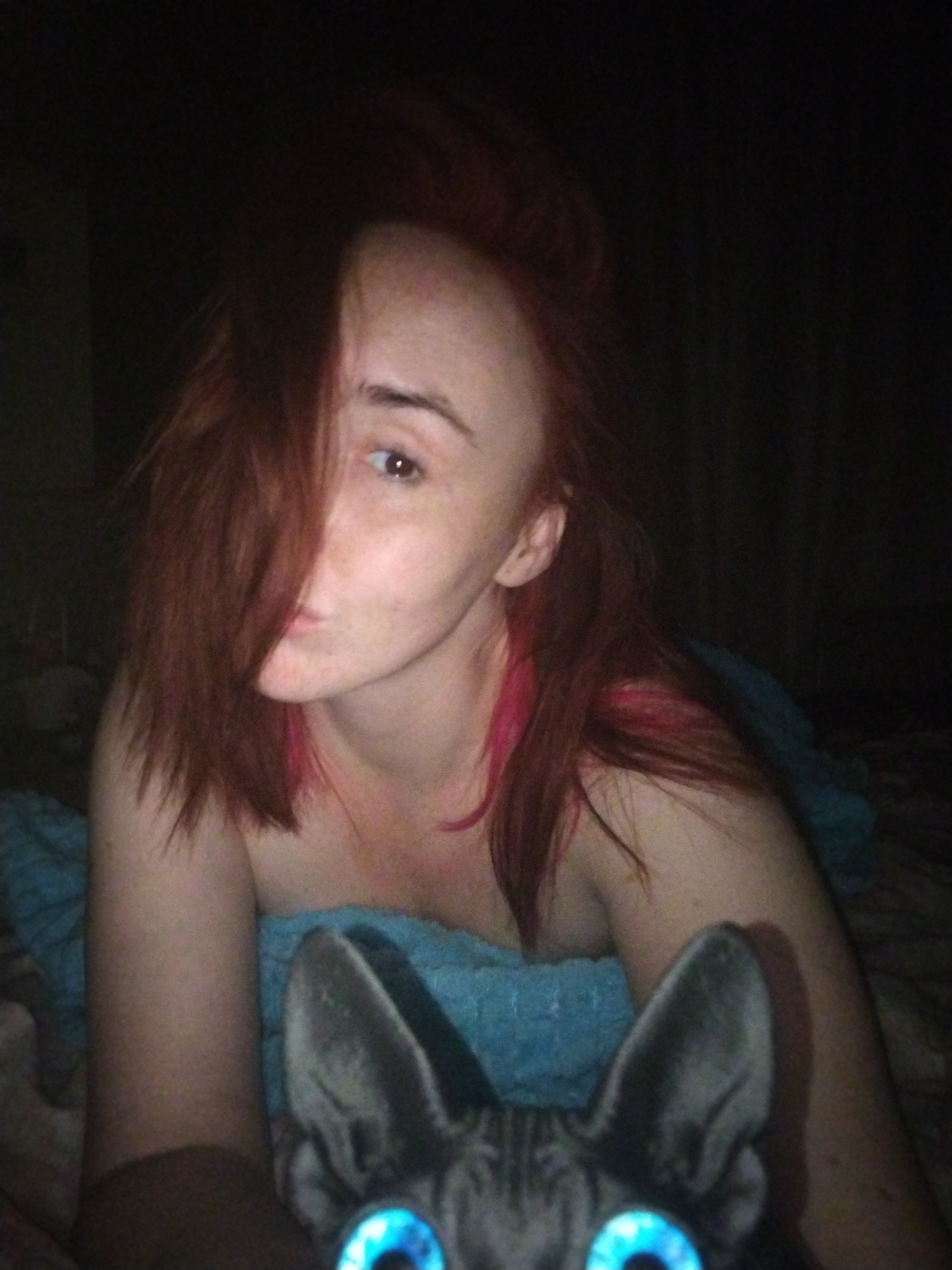 A person with red hair partially obscuring their face is shown in a low-light indoor setting, possibly a bedroom. They are wrapped in a light blue blanket. At the bottom of the frame, a dark cat with striking blue eyes is visible. No information indicates this image was taken in Izhevsk, Russia.