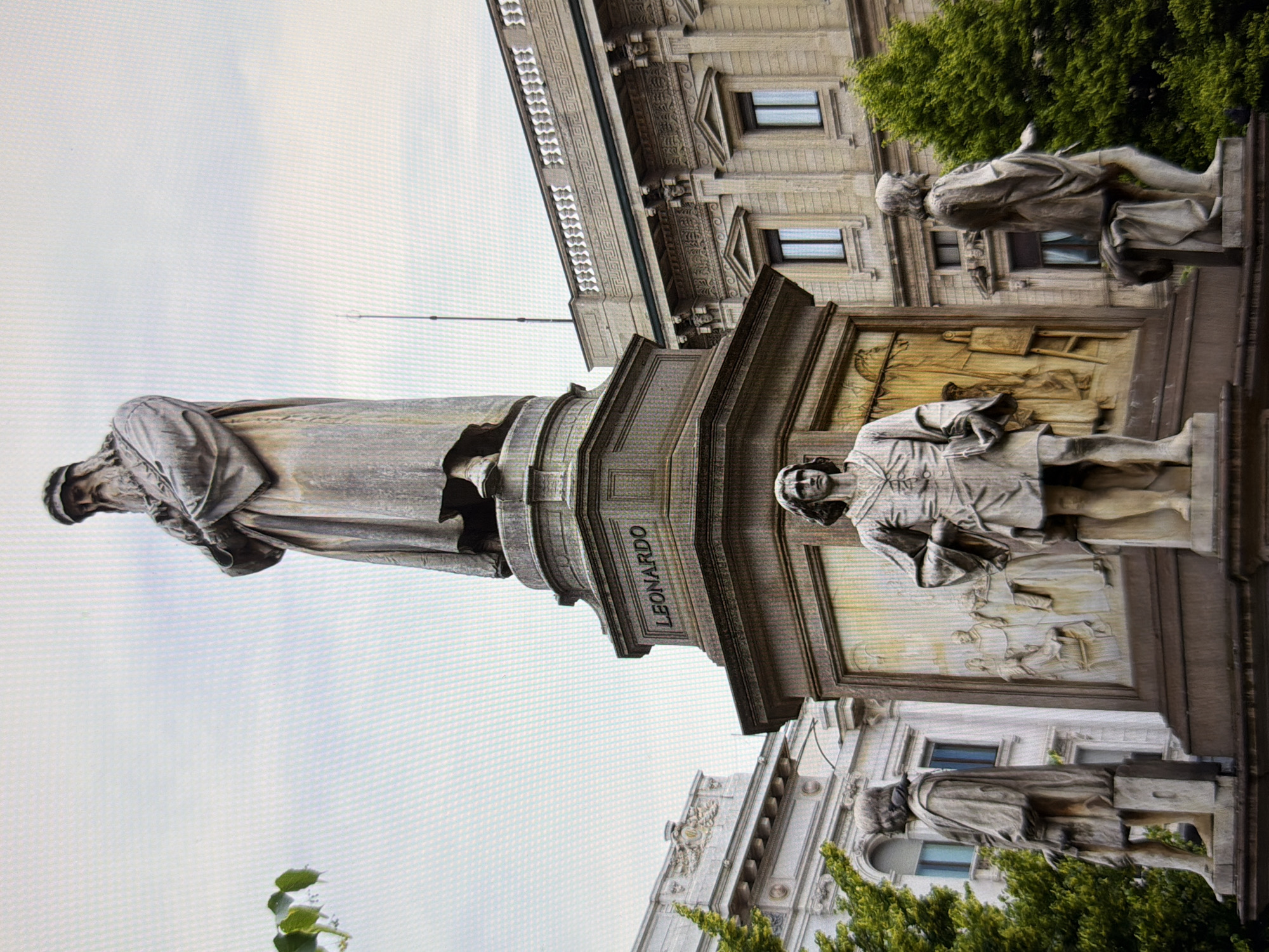 The image shows the Monument to Leonardo da Vinci, located in Piazza della Scala, Milan, Italy. The monument features a large central statue of Leonardo da Vinci in a contemplative pose, with his arms crossed and a beard. He is wearing a long robe. Surrounding the base of the central statue are four smaller statues of his pupils. The monument is made of stone and stands on a large pedestal. In the background, there is a building with classical architectural features, and some trees. The sky is overcast.