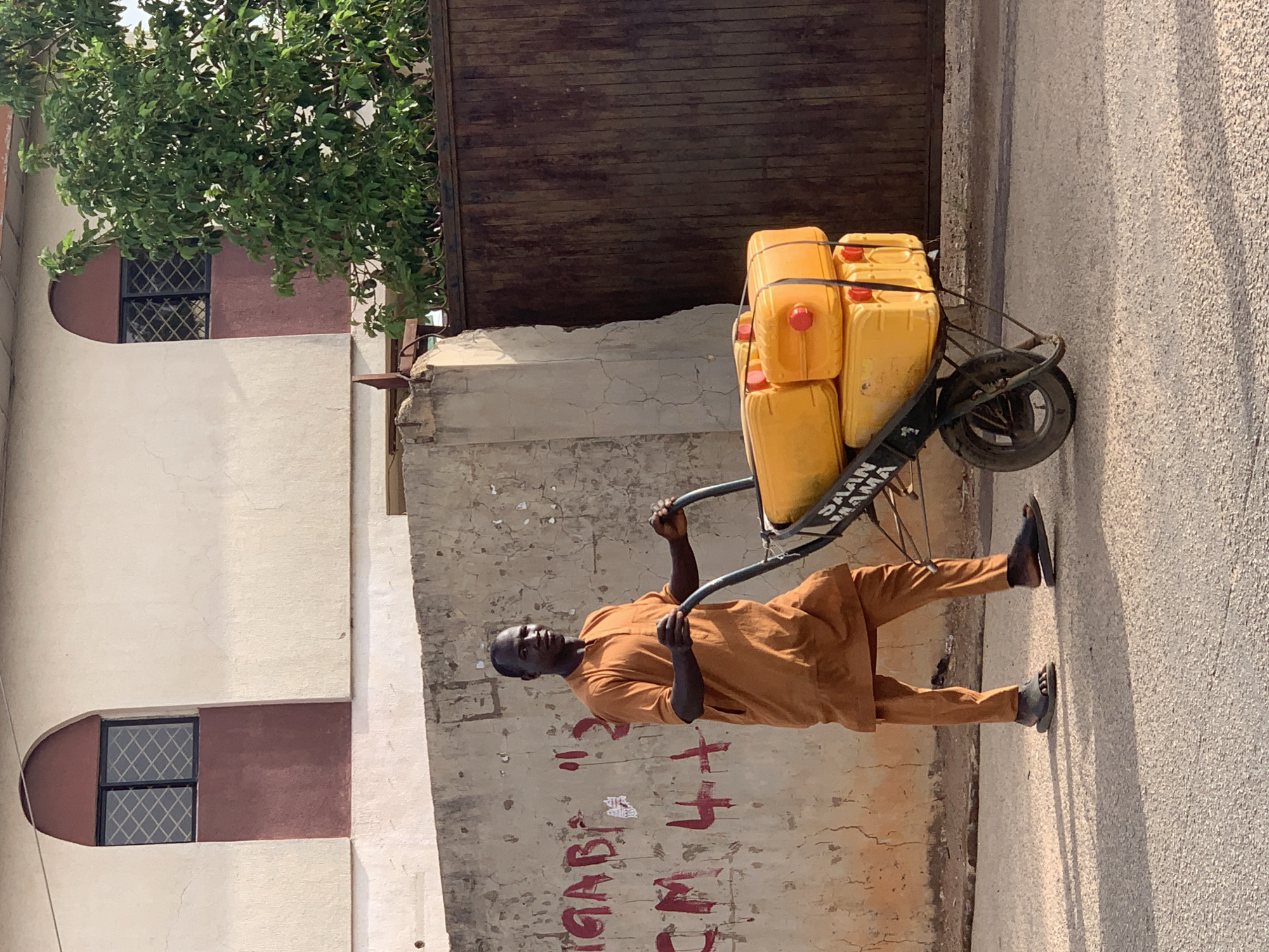 A man in an ochre-colored tunic and trousers pushes a heavily laden wheelbarrow in Jos, Nigeria. The wheelbarrow holds several yellow jerrycans, secured with black straps. The man wears dark sandals. The backdrop includes a weathered wall covered in faded graffiti, a tree, a building with arched windows, and a corrugated metal gate. The scene is bathed in the warm light of early morning or late afternoon.