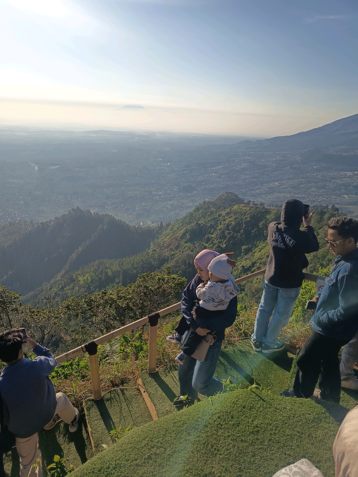 The image captures a scenic overlook in the city of Keditan, Indonesia, with a group of people enjoying the view. In the foreground, a woman wearing a pink hijab holds a baby dressed in a light blue outfit. To her right, a person in a black hooded jacket with white lettering on the back is taking a photo with a camera. Further to the right, a man in a denim jacket and sunglasses is looking at his phone. On the left side of the frame, a person in a blue sweatshirt and light-colored pants is also taking a photo with their phone. The group is standing on a wooden railing that separates them from a steep, green hillside. The background reveals a sprawling landscape of lush green hills and a valley dotted with buildings, all under a hazy, bright sky. The overall impression is one of a tourist destination offering a panoramic view of the surrounding natural beauty.