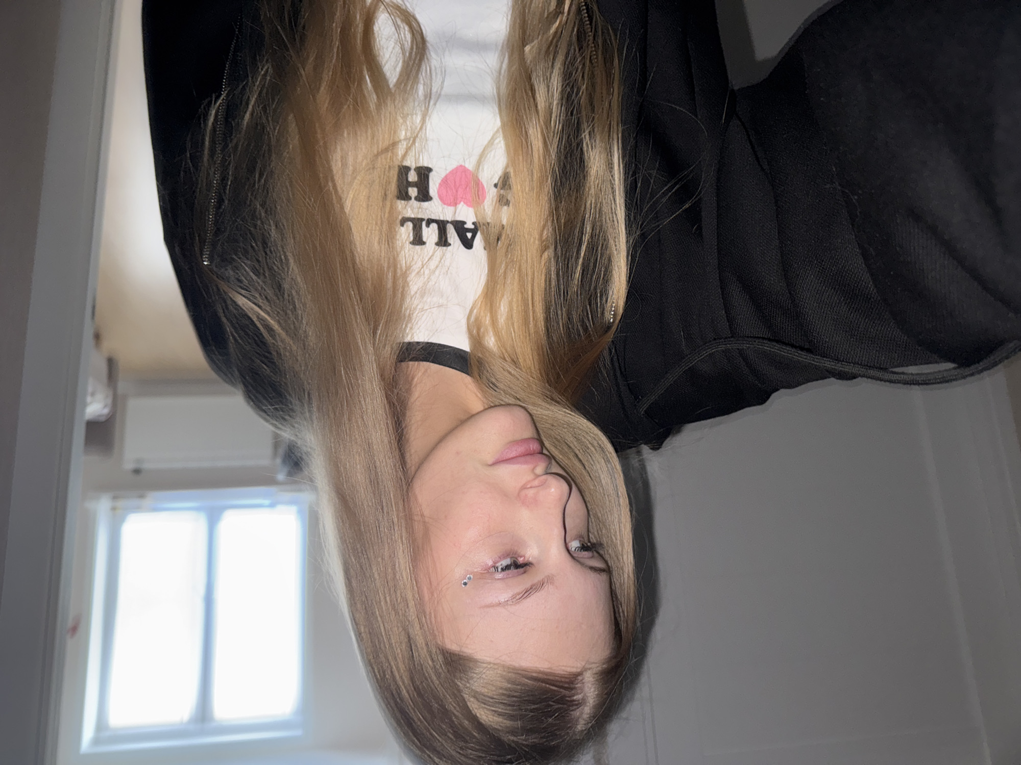 The image shows a young woman with long blonde hair and a black jacket. She is wearing a white t-shirt with black trim and text that appears to say "ALL I H". She has two small, clear rhinestones applied to her left eyebrow. The background is out of focus but appears to be an indoor setting with a window on the left side of the frame. The image is oriented sideways, with the woman's head on the left side of the frame. There is no discernible information about the city of Perm, Russia, or any specific urban location.