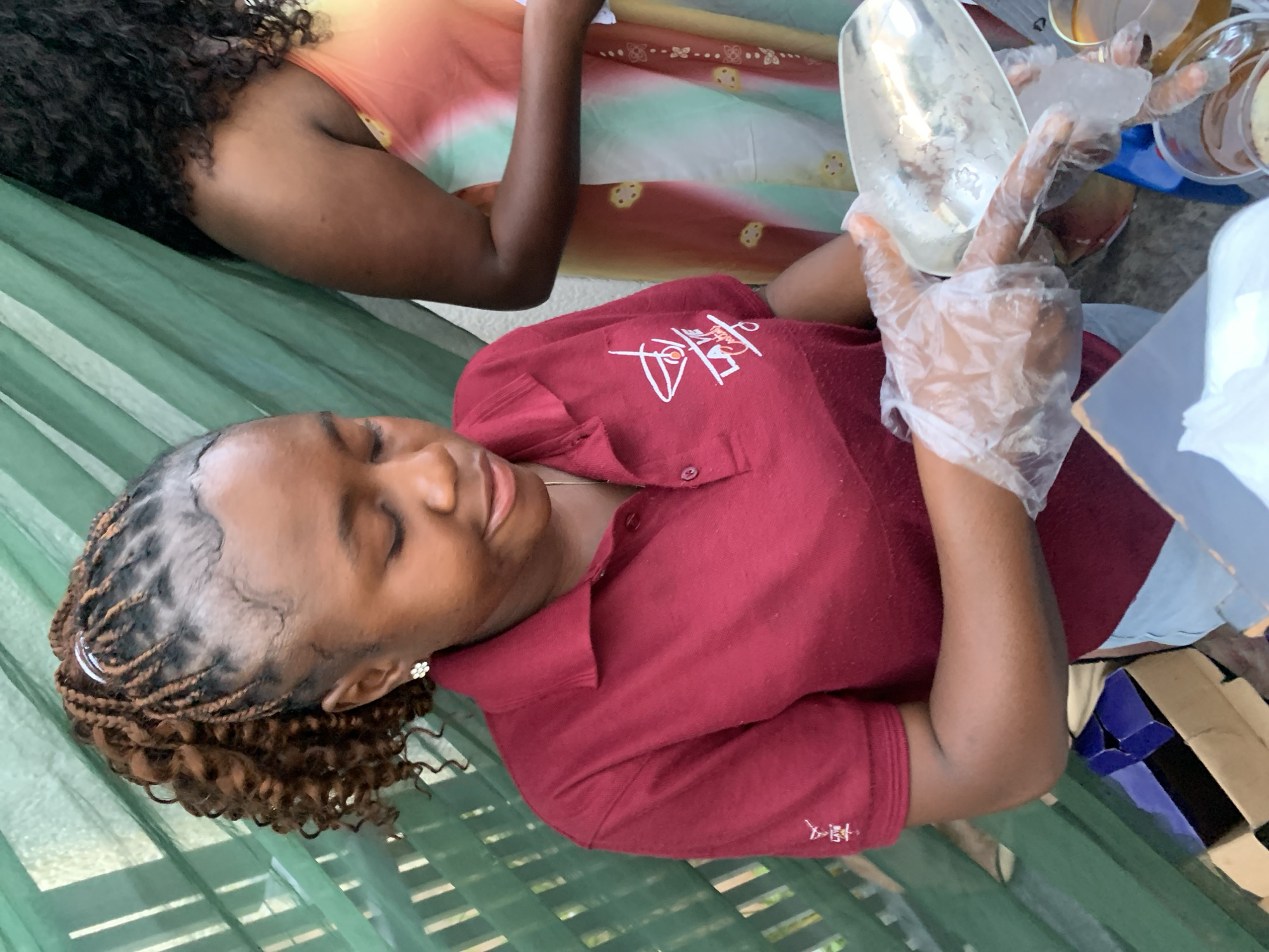 A young Black woman with braided hair and a white flower earring, wearing a maroon polo shirt with a logo on the left chest, is scooping ice into a clear plastic container with gloved hands. She is looking down at her task. Behind her, another Black woman with curly hair is visible, wearing a colorful tie-dye dress. They appear to be outdoors, with a green lattice-like structure in the background. Boxes and cups are present on a surface in the foreground, suggesting a food or drink service setup. The scene takes place in Gura Top, Nigeria.