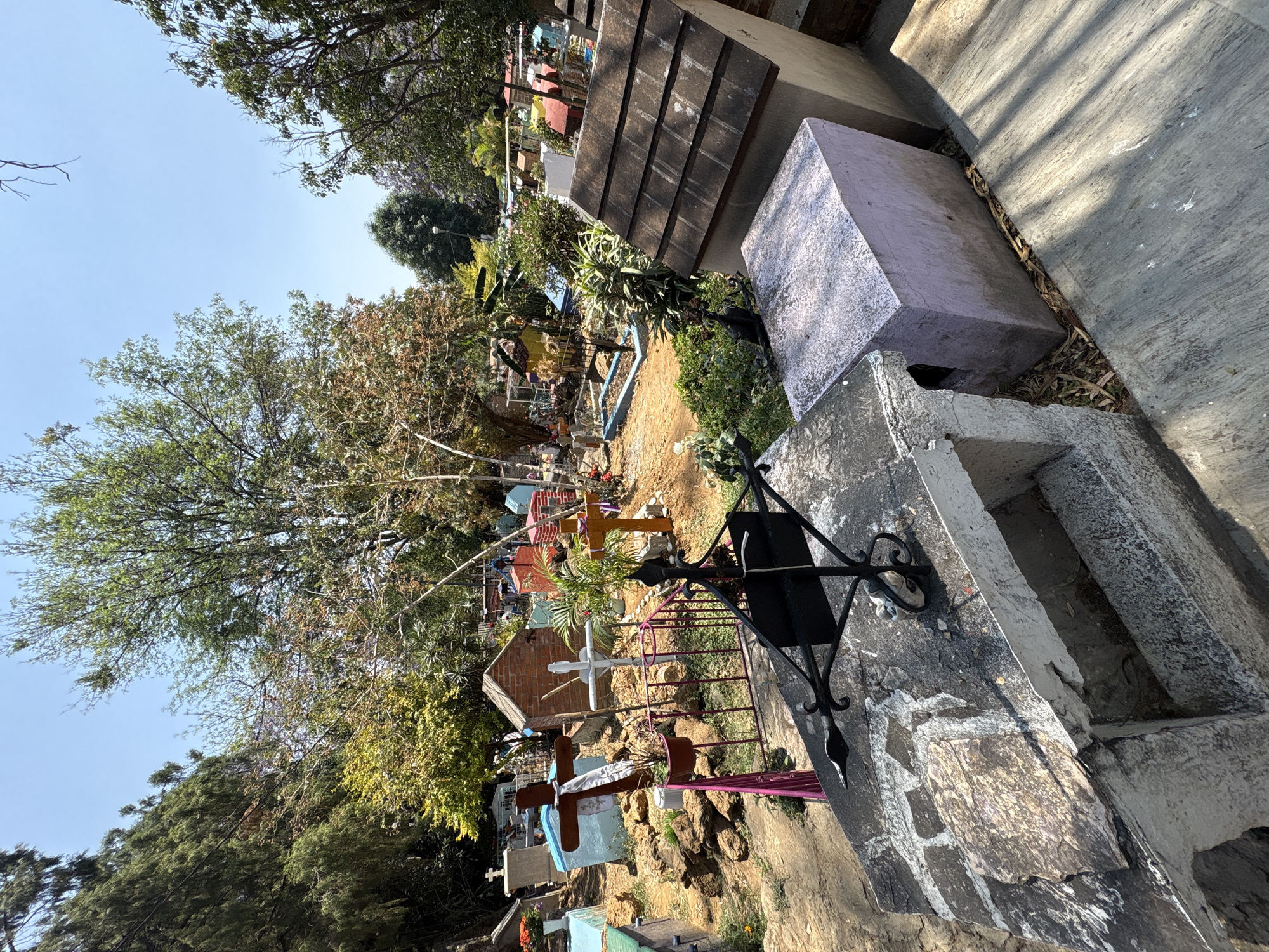 The image depicts a cemetery in Ocotepec, Mexico. The scene is filled with gravestones and crosses of various sizes and colors, amidst trees and vegetation. Some graves are decorated with flowers and other offerings. The foreground features a weathered concrete structure with a metal cross. The overall atmosphere is serene and contemplative.