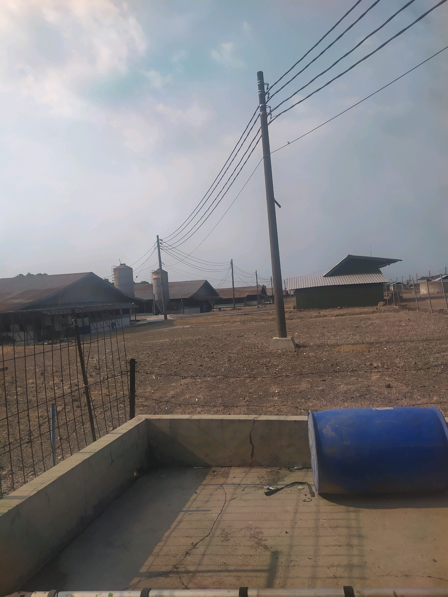 This is a wide shot of a rural or semi-rural outdoor scene, possibly a farm or agricultural facility in Winong, Indonesia. In the foreground, there is a concrete structure and a large blue barrel lying on its side. A wire fence separates this area from the rest of the scene. In the midground, there are several large, dark-colored buildings with sloped roofs, possibly barns or storage facilities. Two tall, cylindrical silos are visible behind one of these buildings. Power lines stretch across the sky, supported by a utility pole. The ground appears to be dry and dusty. The sky is mostly overcast with some patches of light blue. There are no people visible in the image.