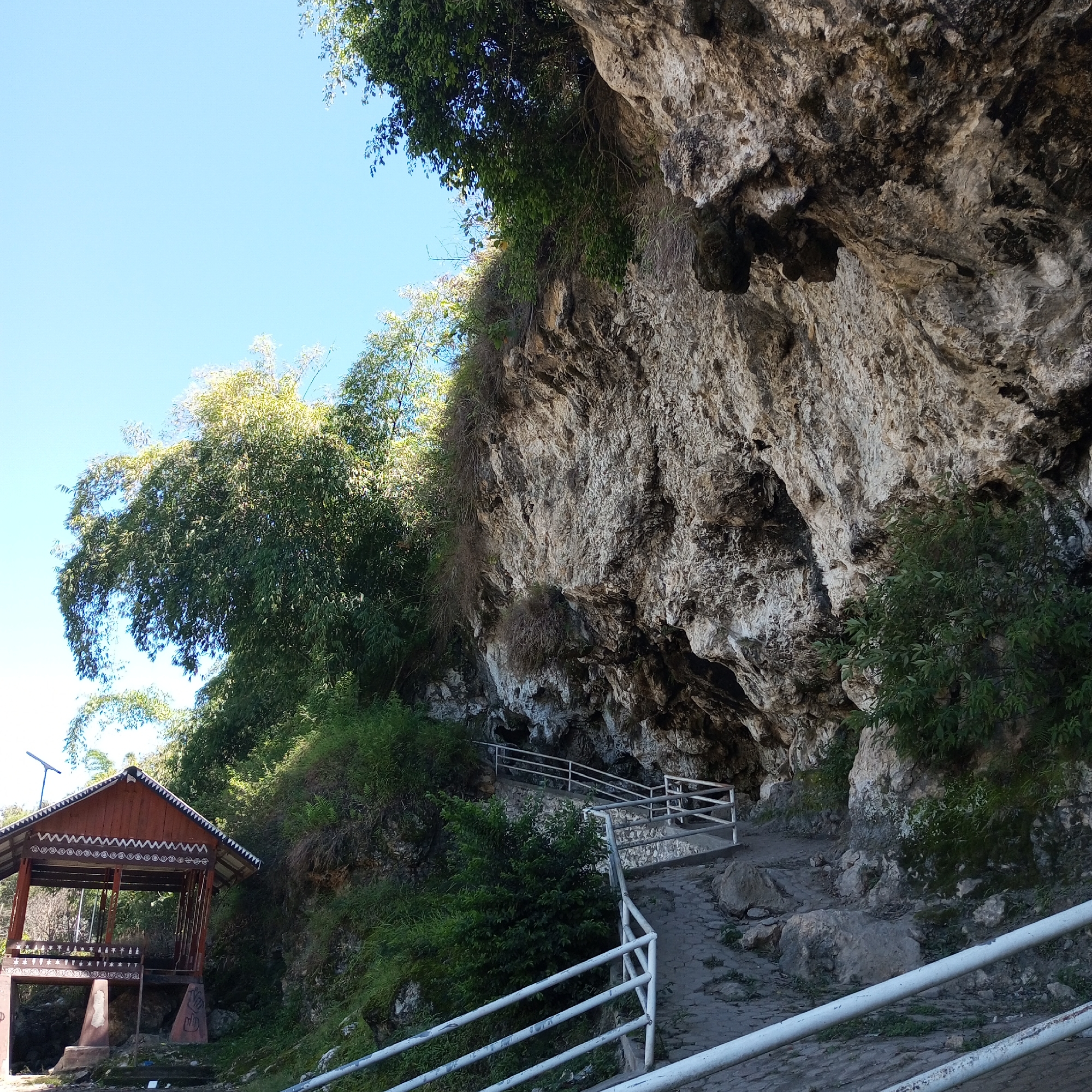 The image depicts an outdoor scene at a natural cave entrance in Kalalengkio, Indonesia. The foreground features a stone pathway with white metal railings, leading towards the cave. The pathway is partially paved with irregular stone slabs and scattered rocks. To the left, a traditional wooden structure with a distinctive patterned roof and white decorative trim stands prominently. This structure appears to be a pavilion or shelter. Lush green vegetation, including trees and bushes, surrounds the pathway and the cave entrance. The cave itself is large and imposing, with a rough, textured rock face that extends upwards. The sky is clear and blue. The scene conveys a sense of natural beauty and potentially a tourist attraction.